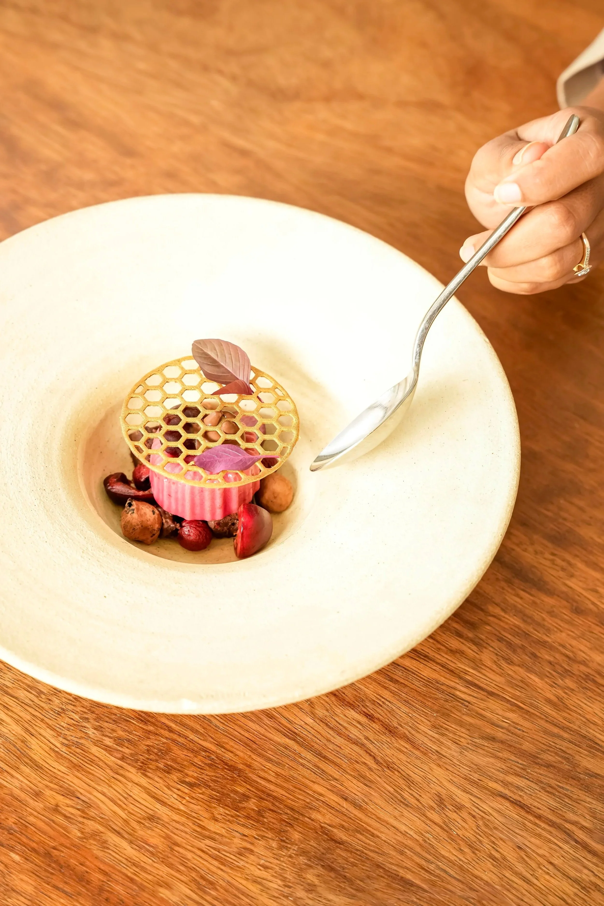 A plated dessert with a pink structured base, a honeycomb design chocolate decoration, small chocolate and berry elements, and purple edible leaves, served in a beige bowl on a wooden table.