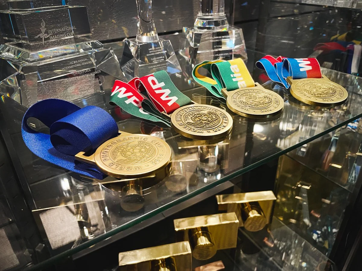 Display of medals and awards, including three gold medals with colorful ribbons and several acrylic trophies in the background, on a glass surface.