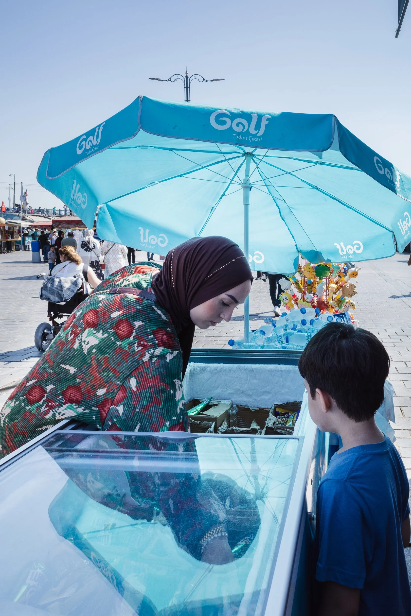 Woman at an ice cream cart leaning toward a boy beneath a large blue umbrella.
