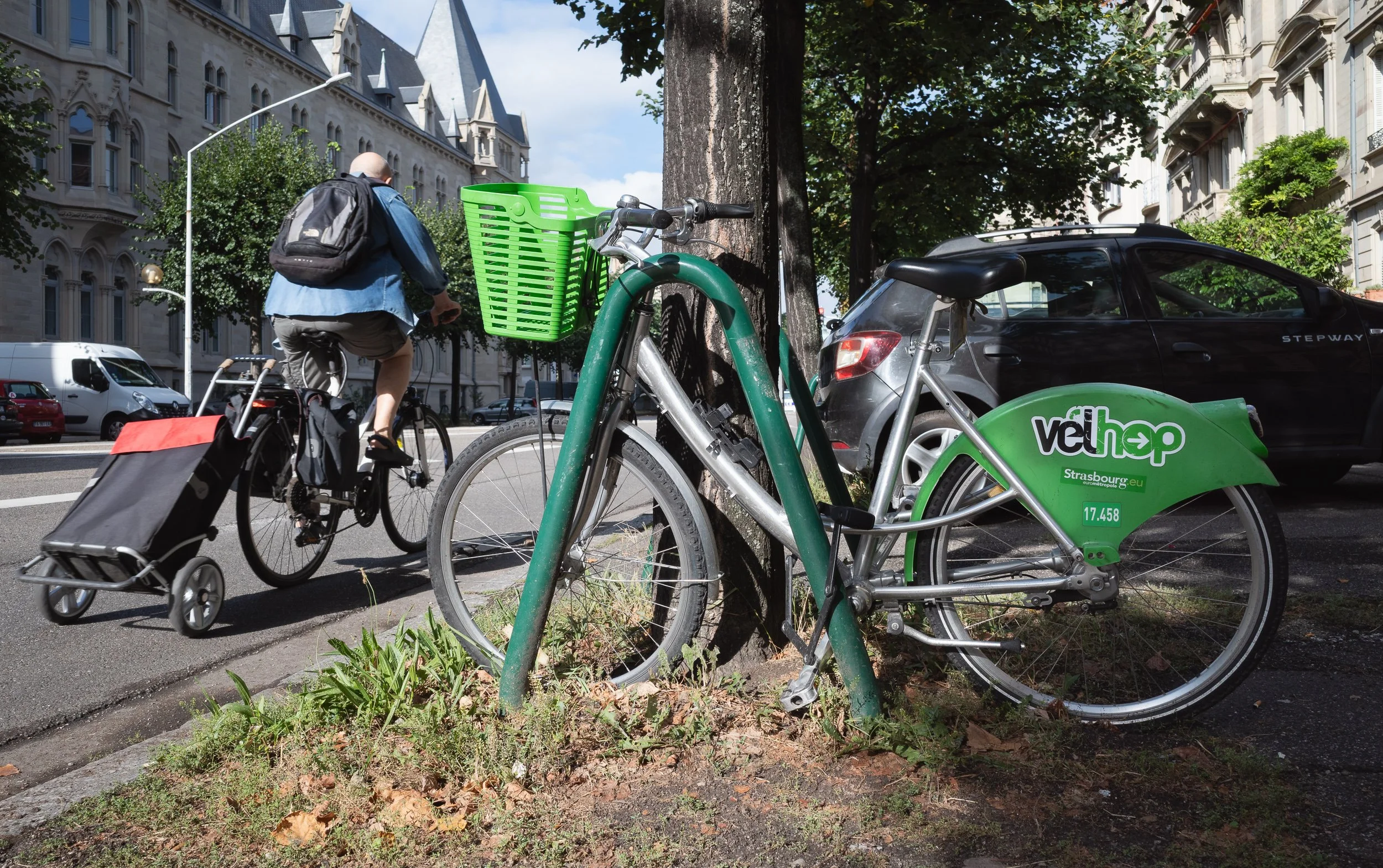 Vélhop bicycle parked by a tree as a cyclist pulls a wheeled load behind another bike.