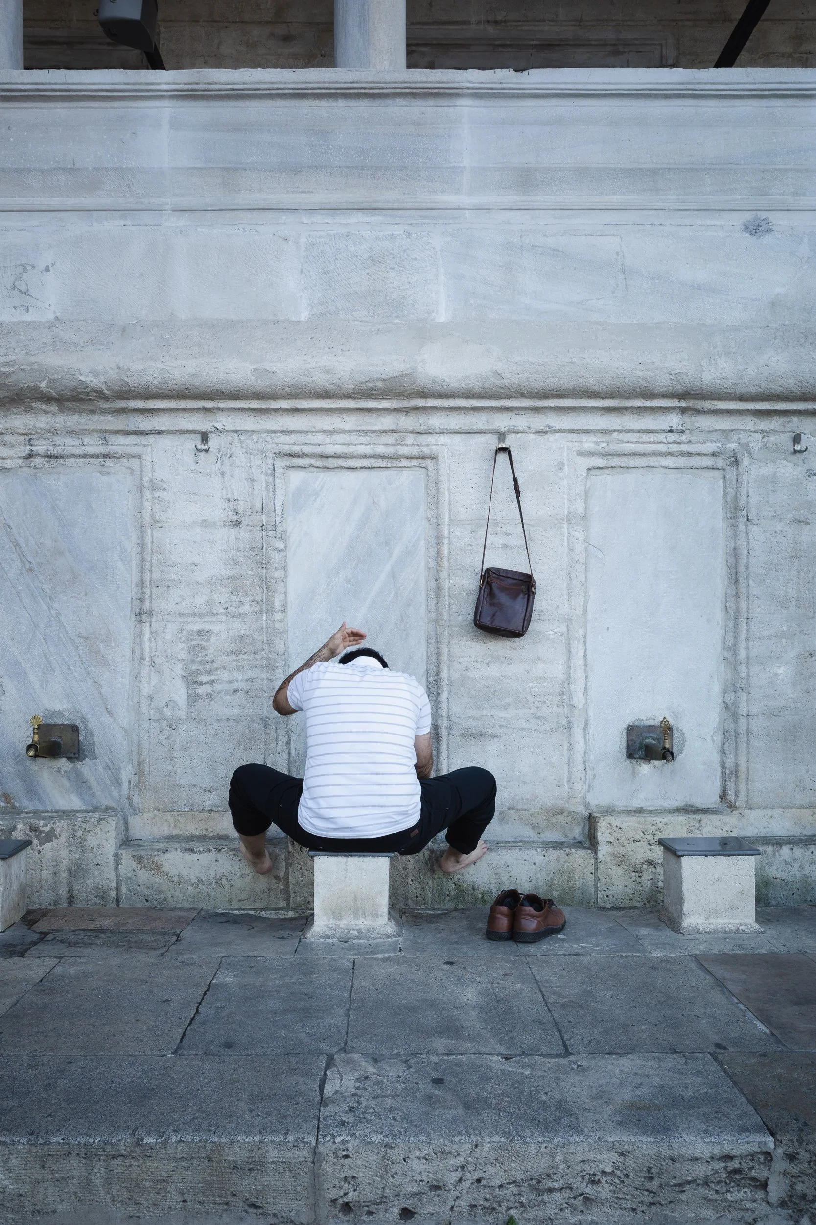 Man sitting against a pale stone wall with his shoes and shoulder bag placed beside him.