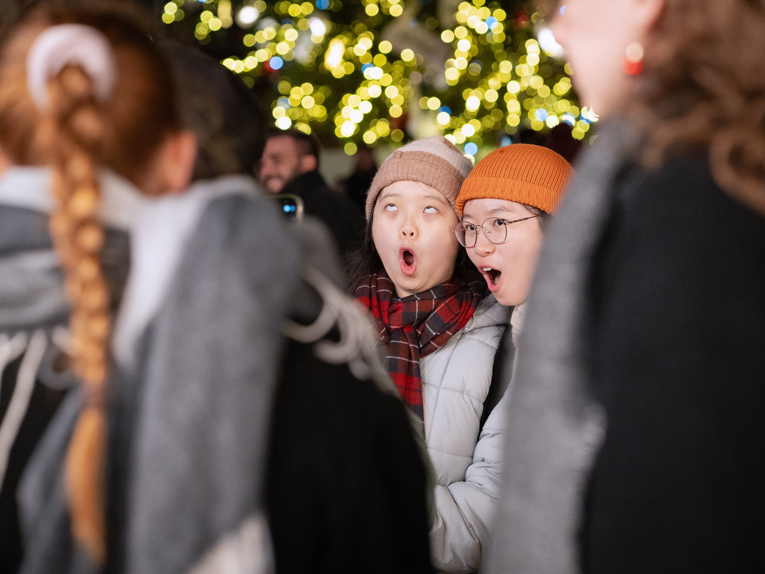 Two young women making surprised expressions at a crowded Strasbourg Christmas market, with festive lights in the background.