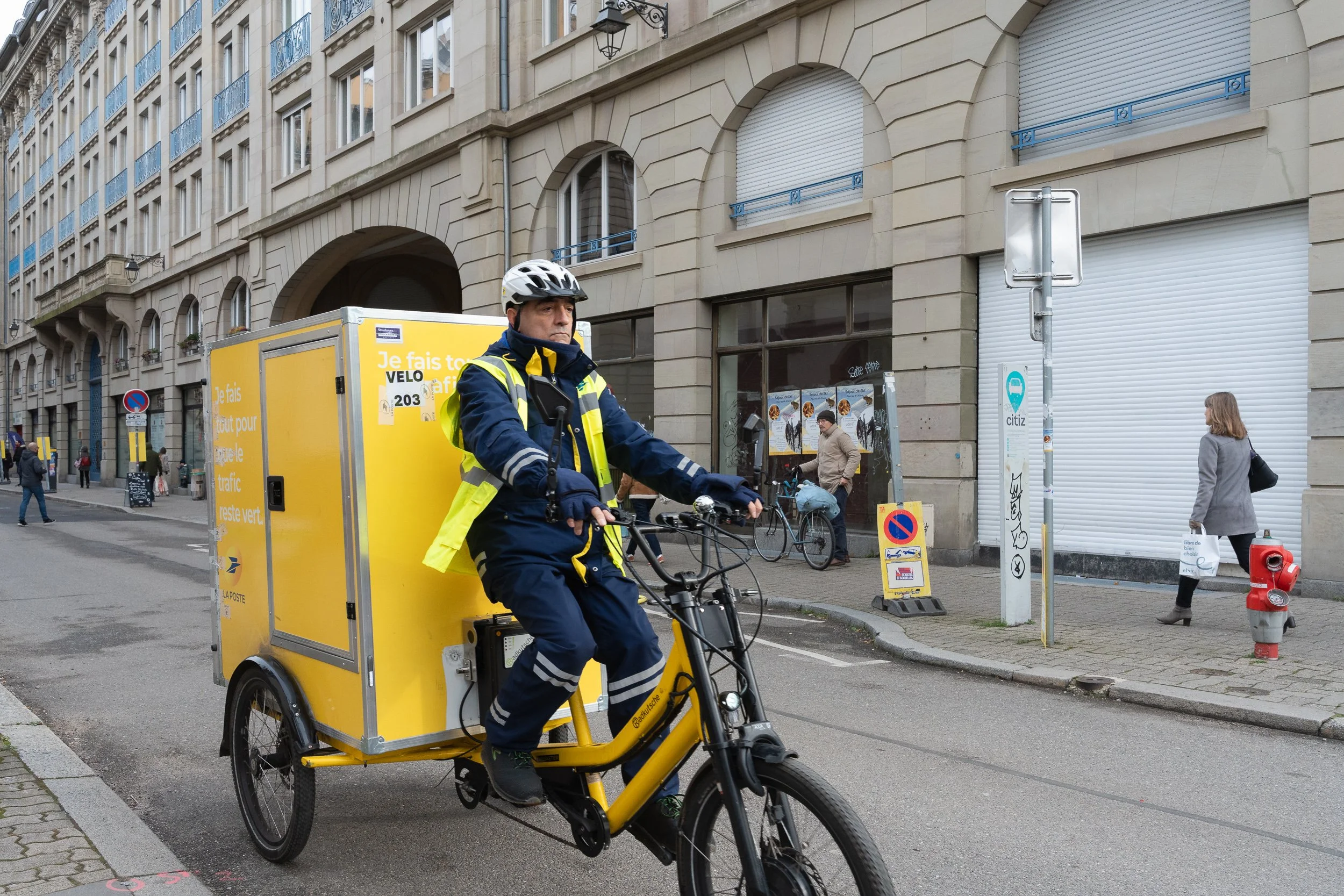 Postal worker riding a yellow cargo bicycle through central Strasbourg.