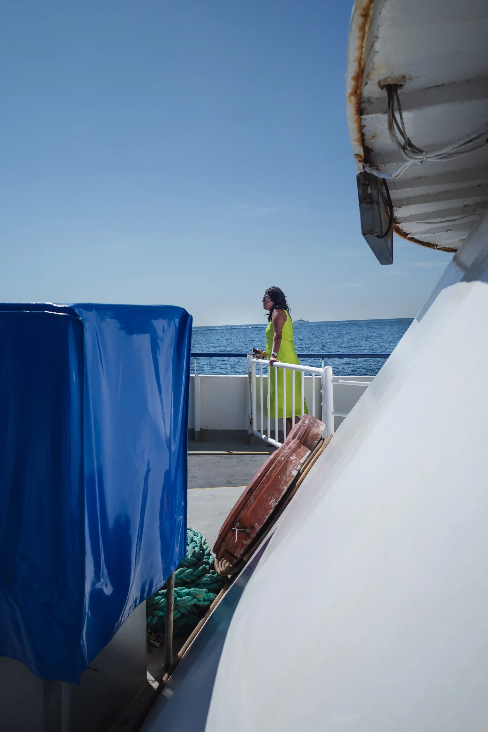 Woman in a bright green dress standing alone on a ferry deck against the sea and blue sky.