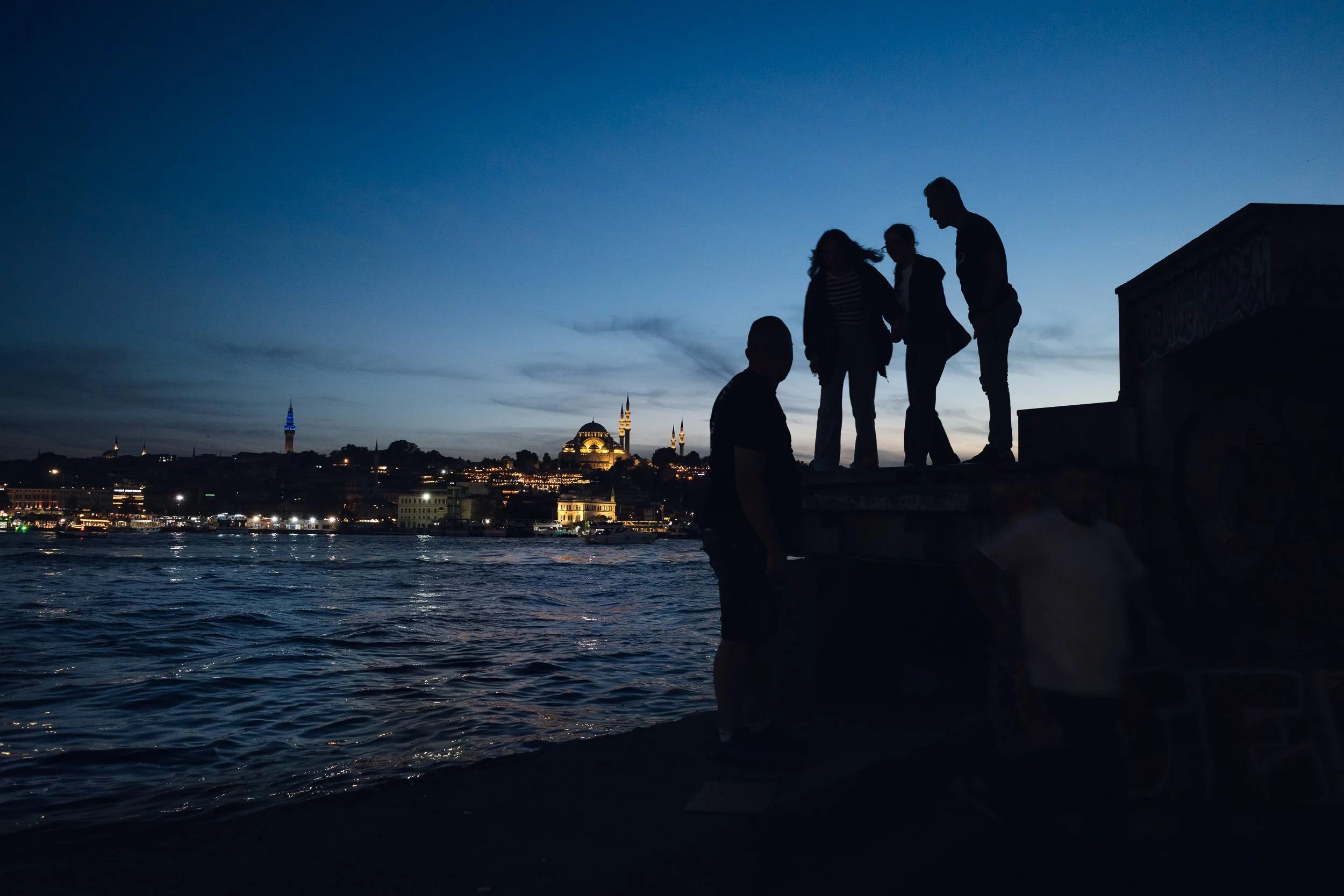 Silhouetted people by the waterfront at dusk with an illuminated Istanbul skyline across the water.