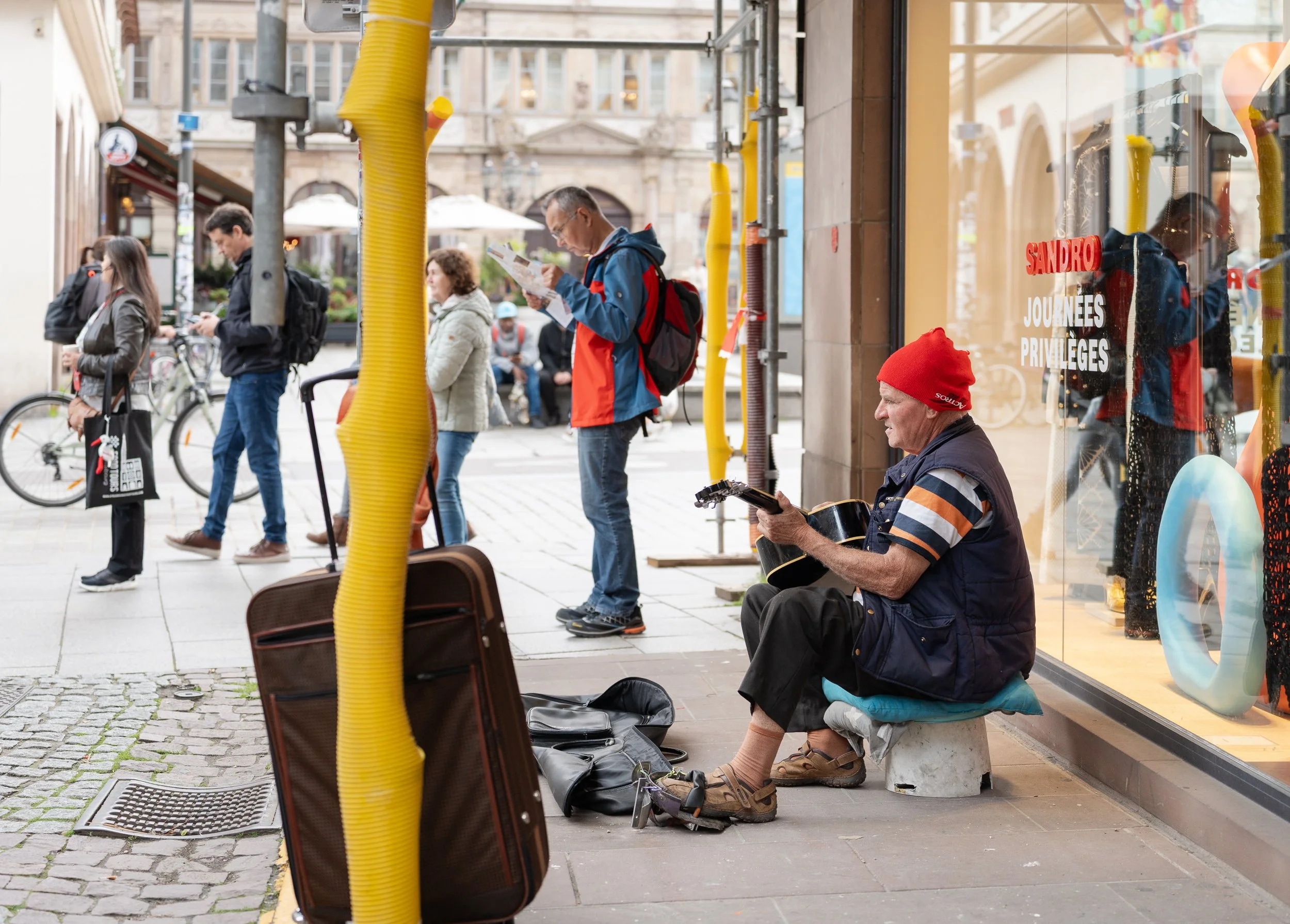 Street musician playing guitar among pedestrians and construction structures in Strasbourg.