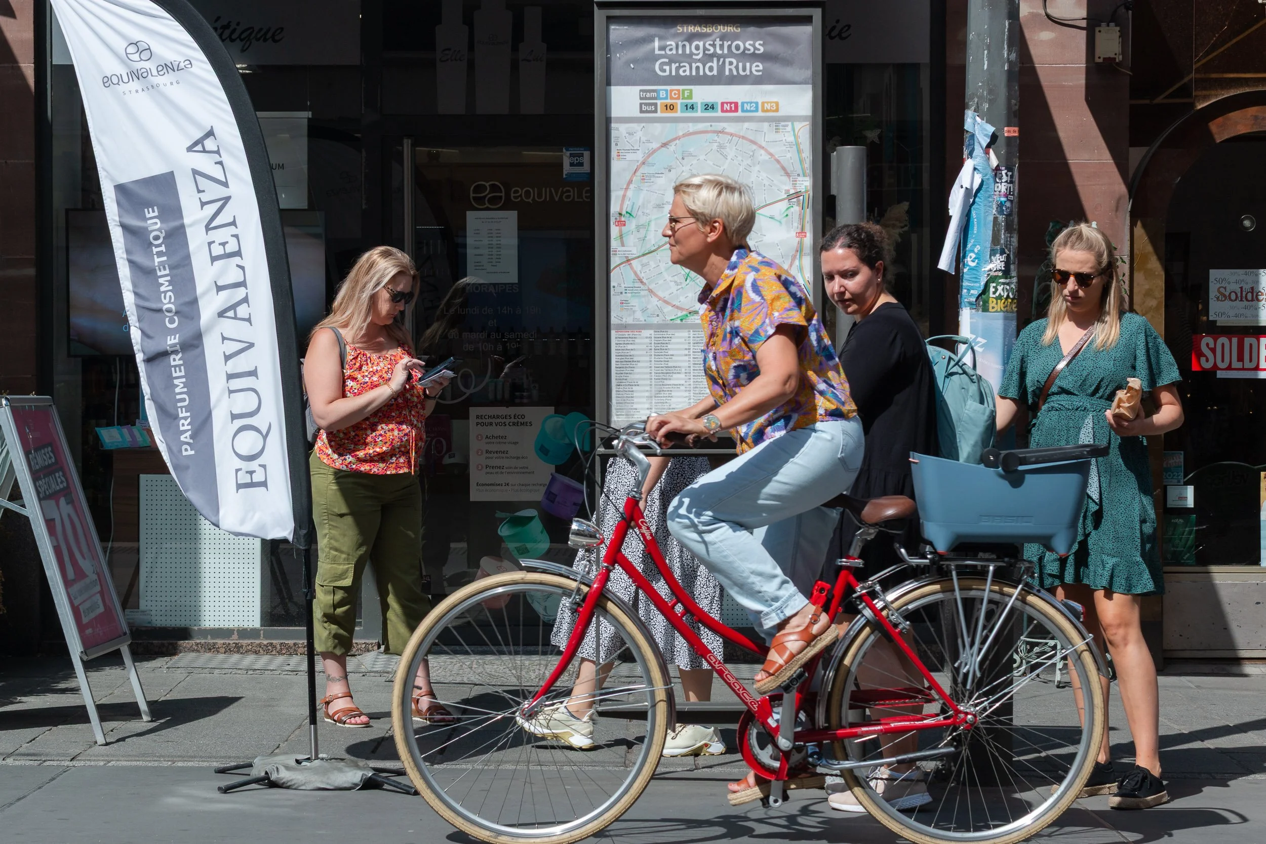 Woman riding a red bicycle past pedestrians and a Strasbourg transport map.