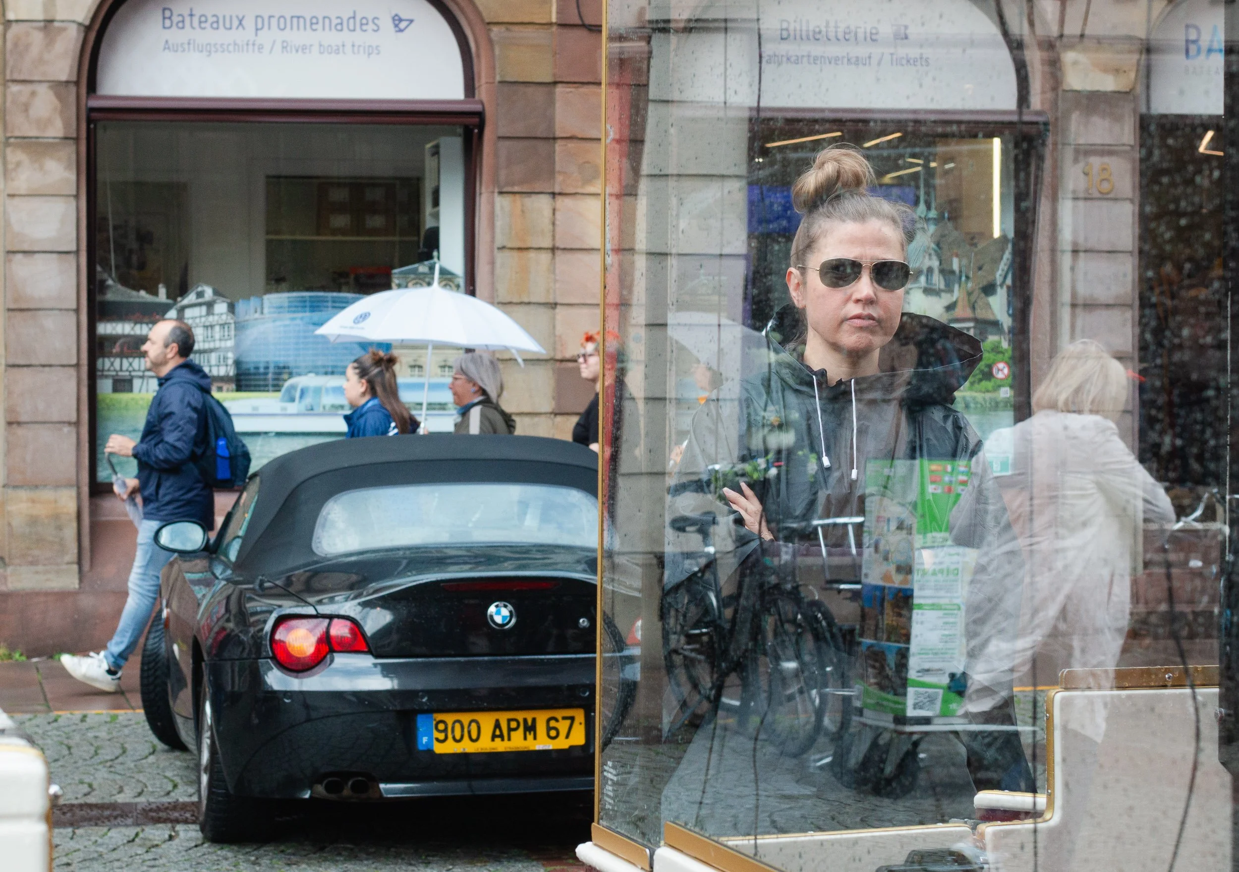 Woman behind glass surrounded by reflections, pedestrians and a parked car.
