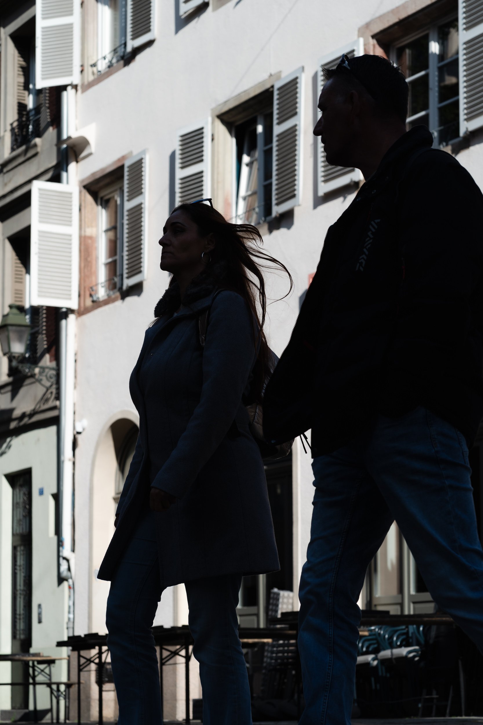 A woman and a man walking through a sunlit Strasbourg street, their figures partly in shadow against traditional building façades.