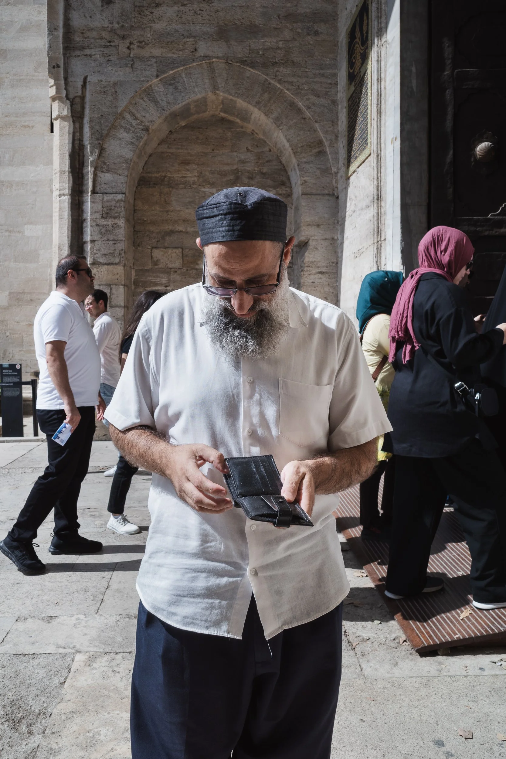 Bearded man in a white shirt looking into his wallet in a stone courtyard in Istanbul.