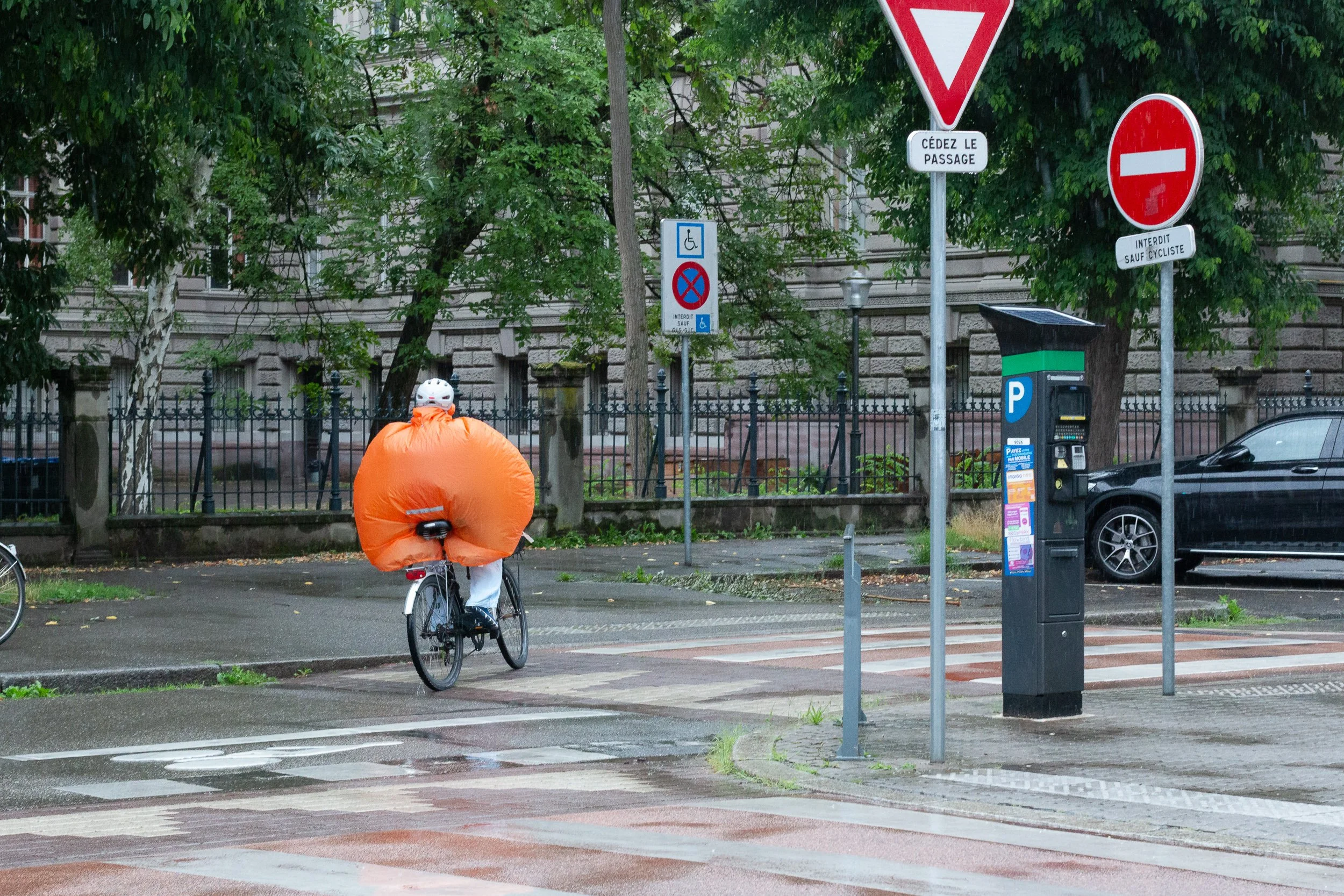 Cyclist wearing a bright orange rain cover on a wet Strasbourg street.