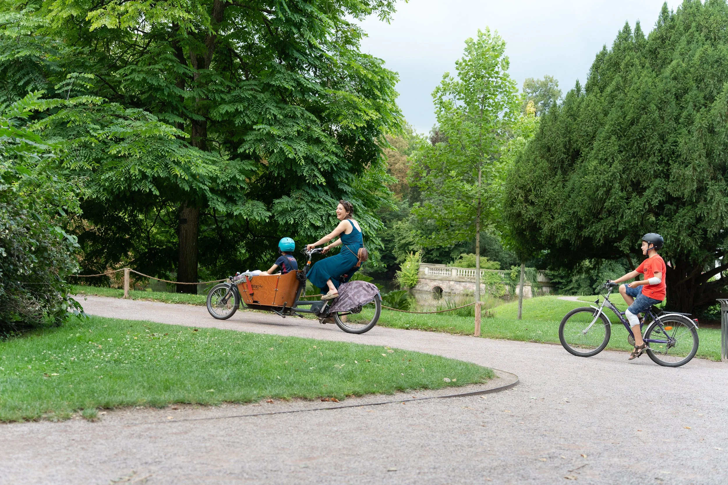 Woman riding a cargo bike with a child while another child cycles beside her in a park.