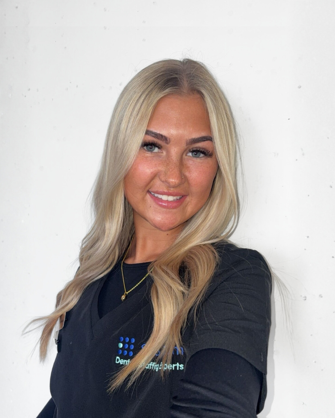 A young woman with long blonde hair, fair skin, and freckles, smiling and wearing a black top with a logo that says "Dental Implant Parts." She is standing against a plain white wall.