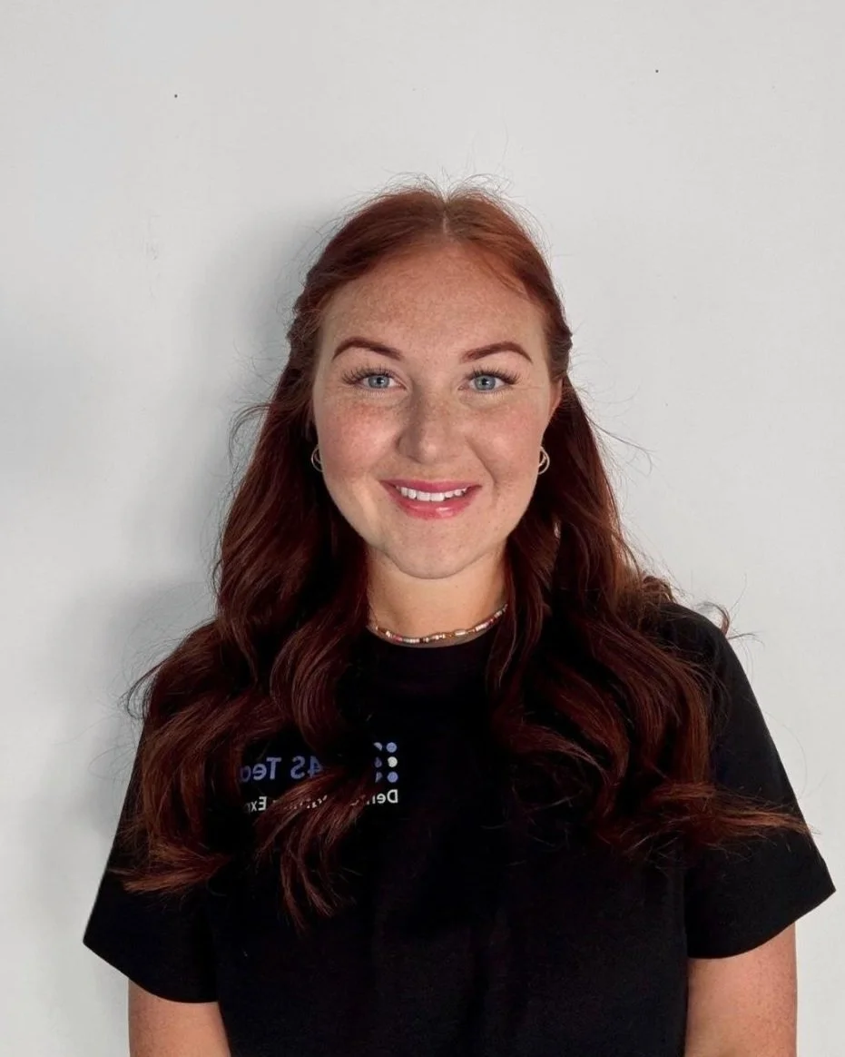 A young woman with long, wavy red hair, blue eyes, and freckles, smiling at the camera against a plain white background, wearing a black T-shirt and a beaded necklace.
