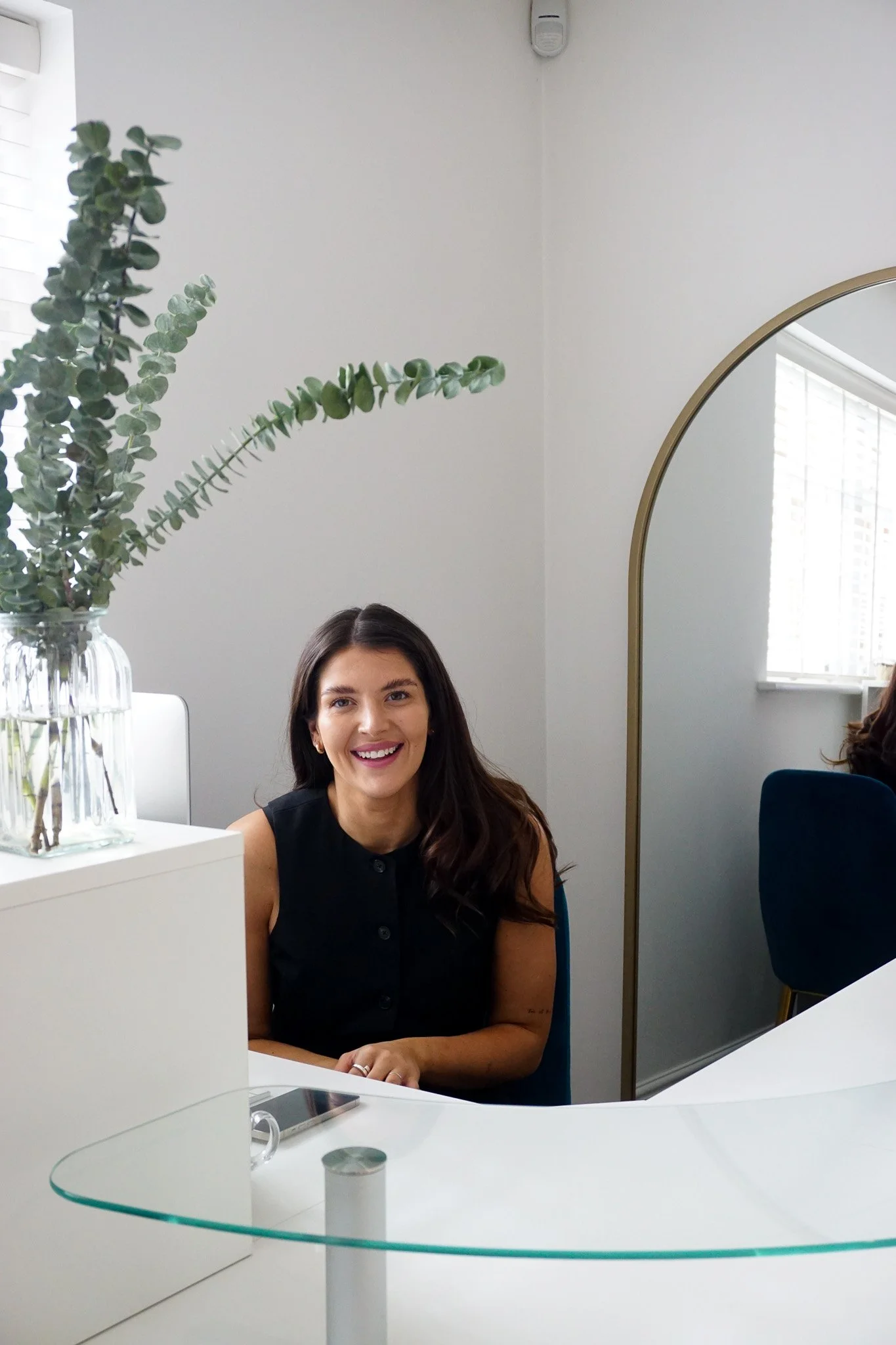 Woman with long dark hair smiling, sitting at a desk in a bright office with a white wall, large window with blinds, and a decorative mirror.