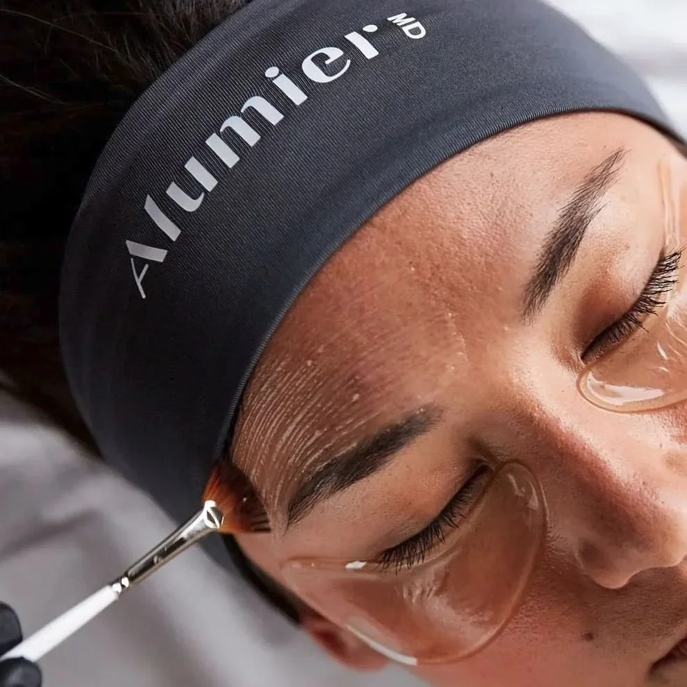 A woman undergoing a skincare treatment, with a skincare brush applying product to her forehead while she rests with eyes closed, wearing a dark headband labeled 'AM Unify' and glasses.