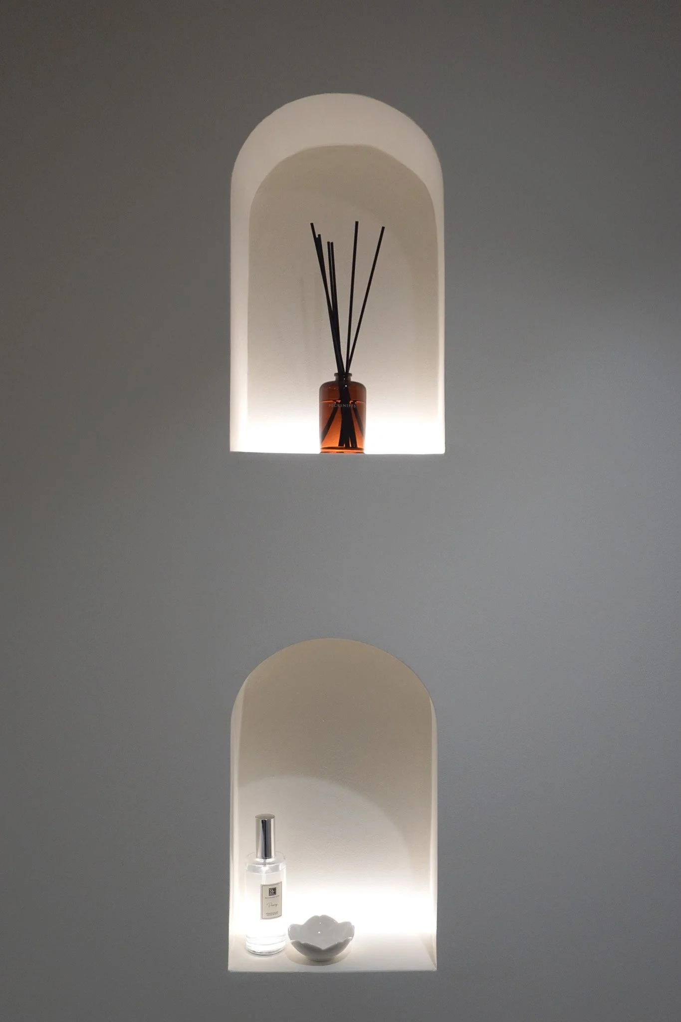 Decorative display niche with a diffuser bottle, a small bowl, and a spray bottle on a bottom shelf, illuminated with built-in lighting.