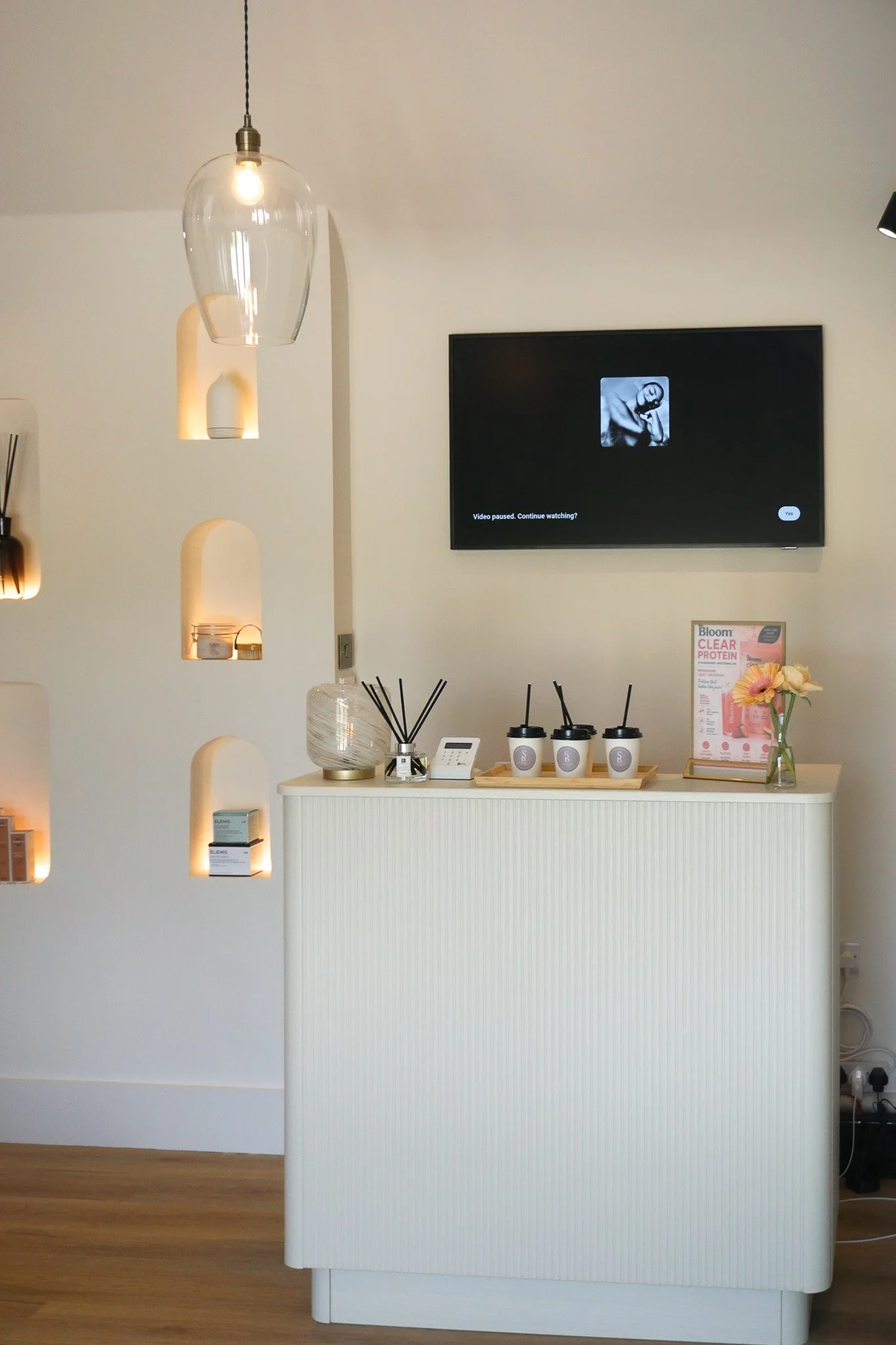 A white reception desk with three black and white coffee cups on a tray, a sign promoting a collagen supplement, a glass vase with pink and orange flowers, a diffuser, and a cordless phone inside a modern, light-filled space.