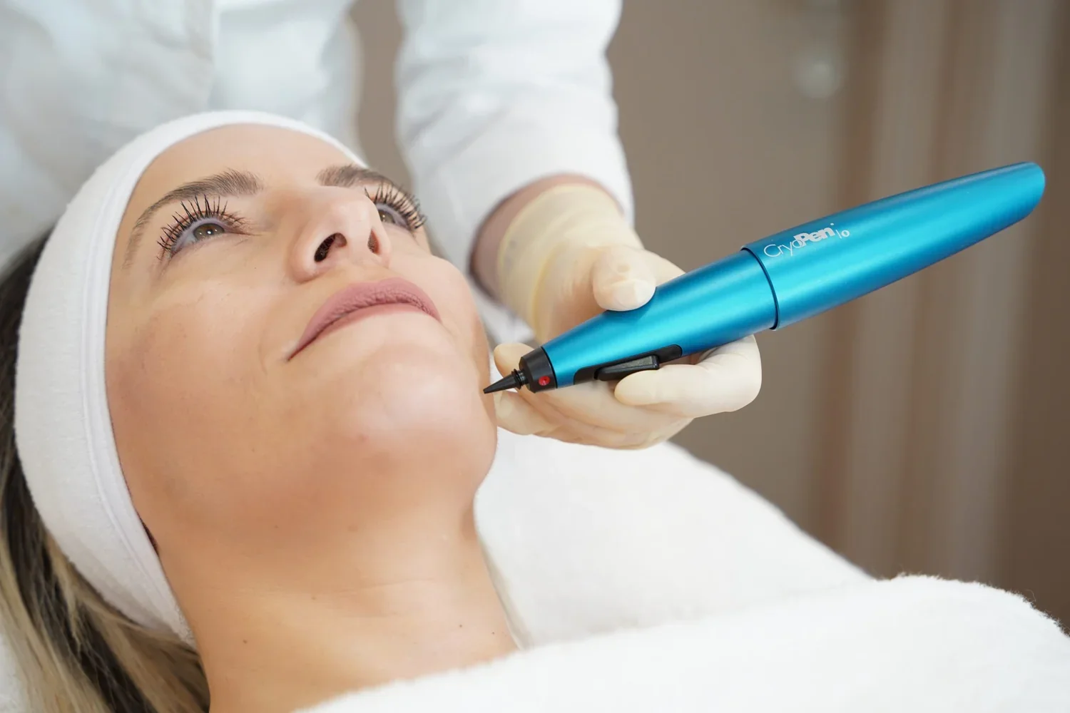 A woman receiving a cosmetic treatment with a blue handheld device on her face, lying down with a white headband and a white gown, in a clinical setting.