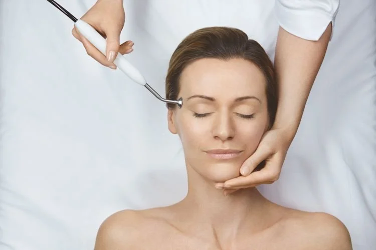 Woman receiving a facial treatment with a handheld device in a spa or clinic setting, lying with eyes closed.
