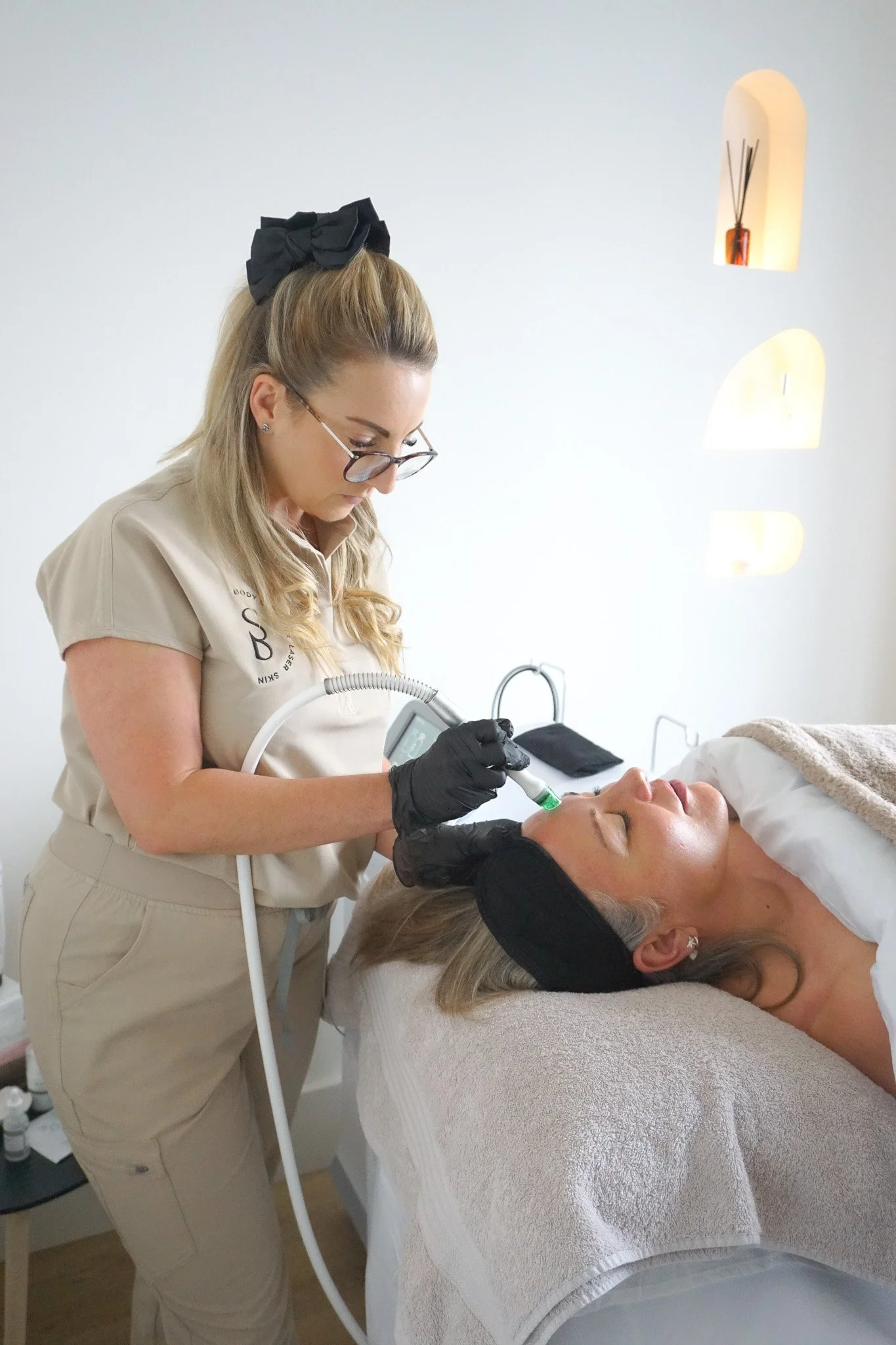 A woman is lying on a treatment bed with her eyes closed, receiving a cosmetic procedure from a professional who is using a handheld device. The professional is wearing a beige uniform, gloves, and glasses, while the woman has a black headband and a 