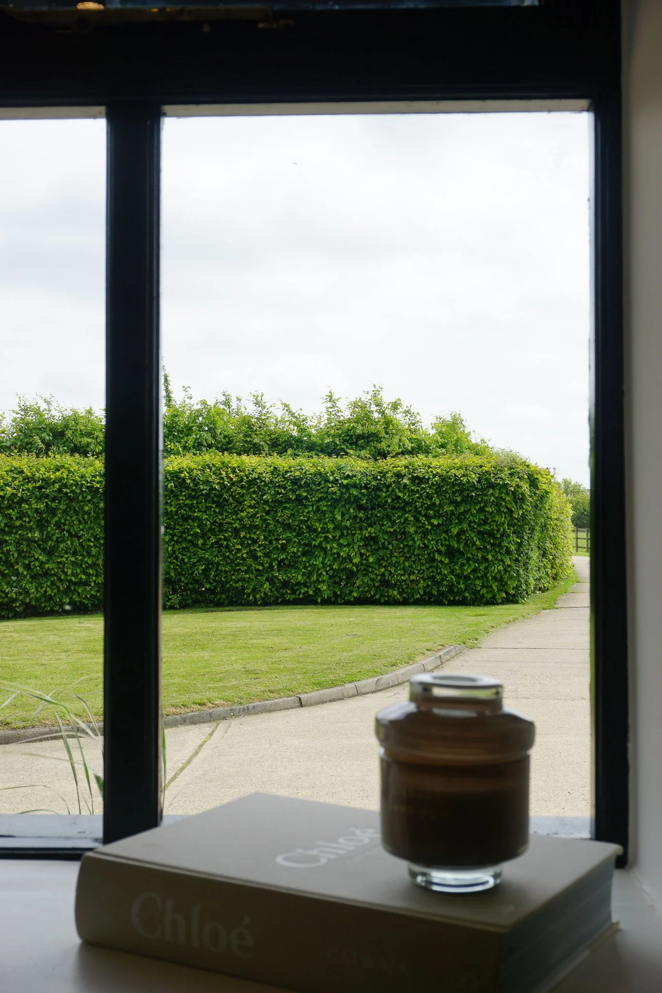 A window looking out onto a well-kept garden with neatly trimmed bushes and a pathway, with a box of chocolates and a small jar placed on a table inside.
