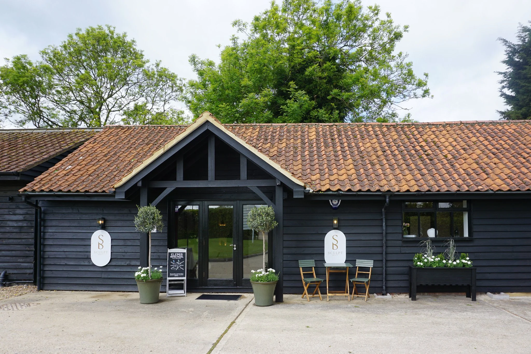 Black wooden building with a pitched roof covered in reddish-brown tiles. The entrance features a glass door, two potted plants, signs with the letter 'S' and 'B', and chairs outside. Large trees with green leaves are visible behind the building.