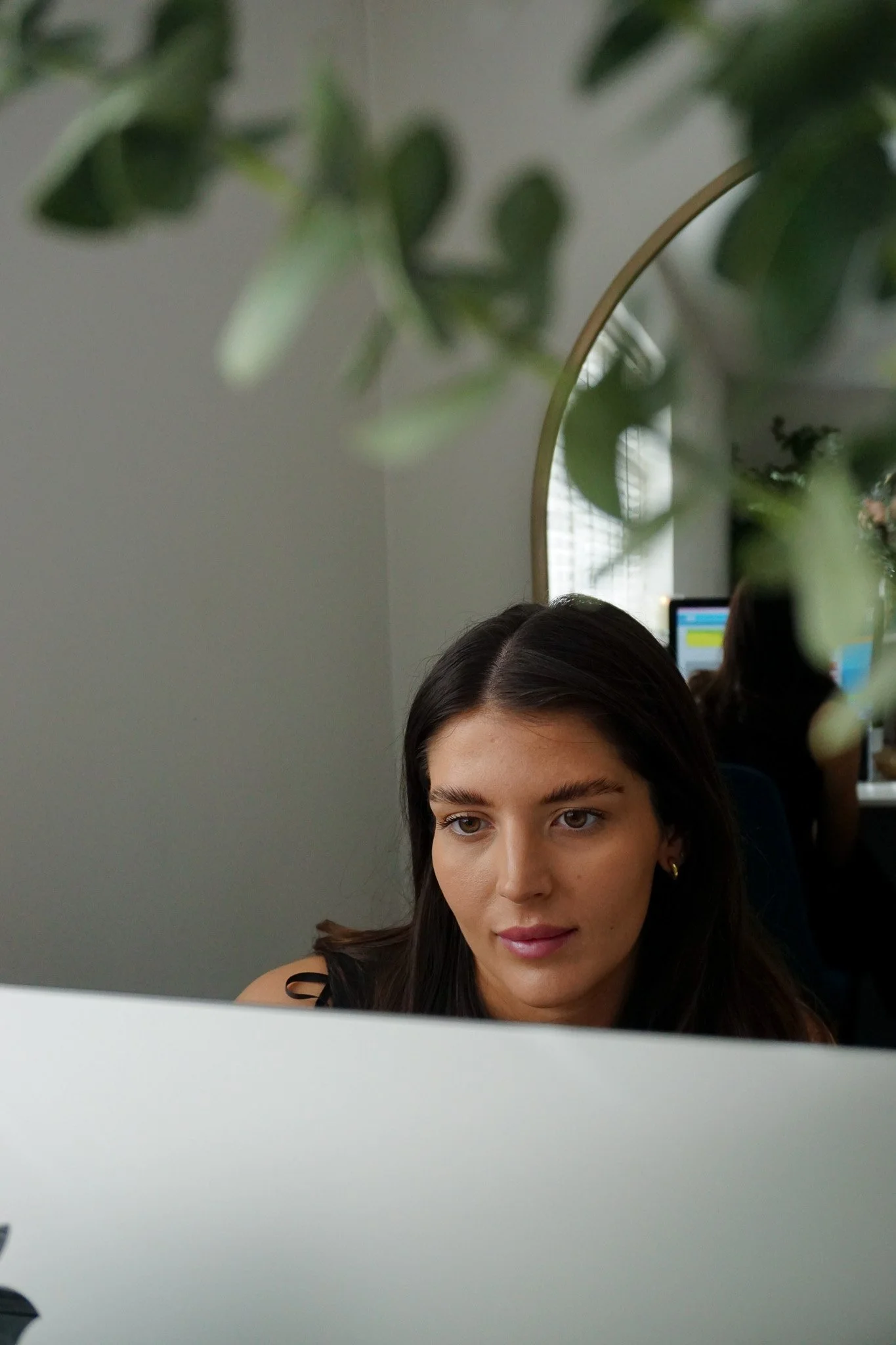 Young woman with long dark hair working at a computer in an office, with greenery and a mirror in the background.
