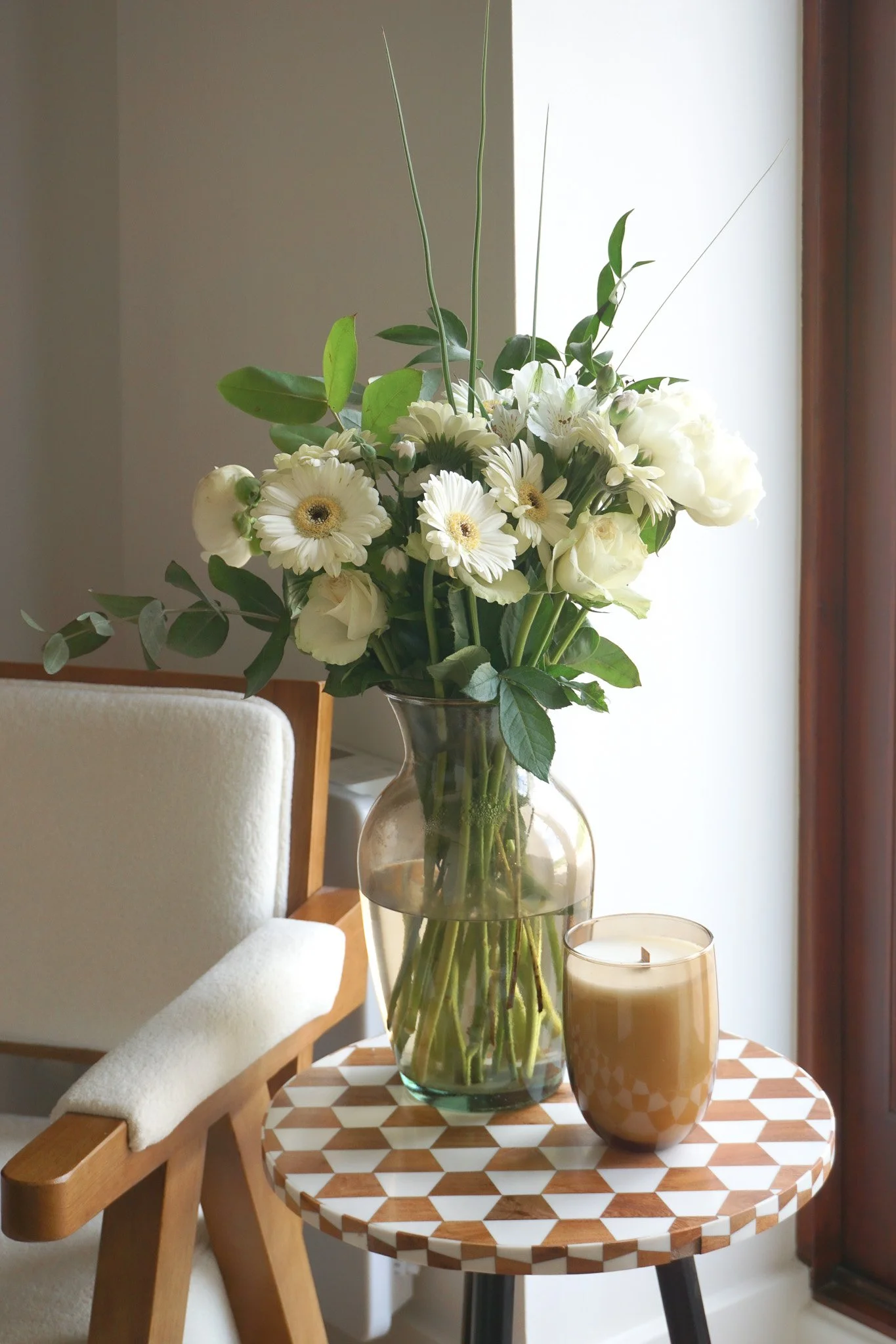 A round wooden table with a geometric pattern on its surface, holding a large glass vase with white flowers and green leaves, and a beige candle in a patterned glass holder. A beige upholstered chair with wooden arms is partially visible on the left 