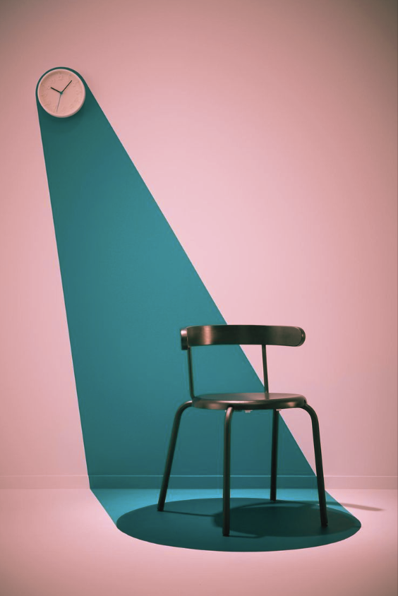 Une chaise noire en métal sous une lumière dirigée créée par une horloge ronde fixée au mur, projetant un rayon de lumière vert clair sur un mur blanc et un sol en bois, dans une pièce moderne.