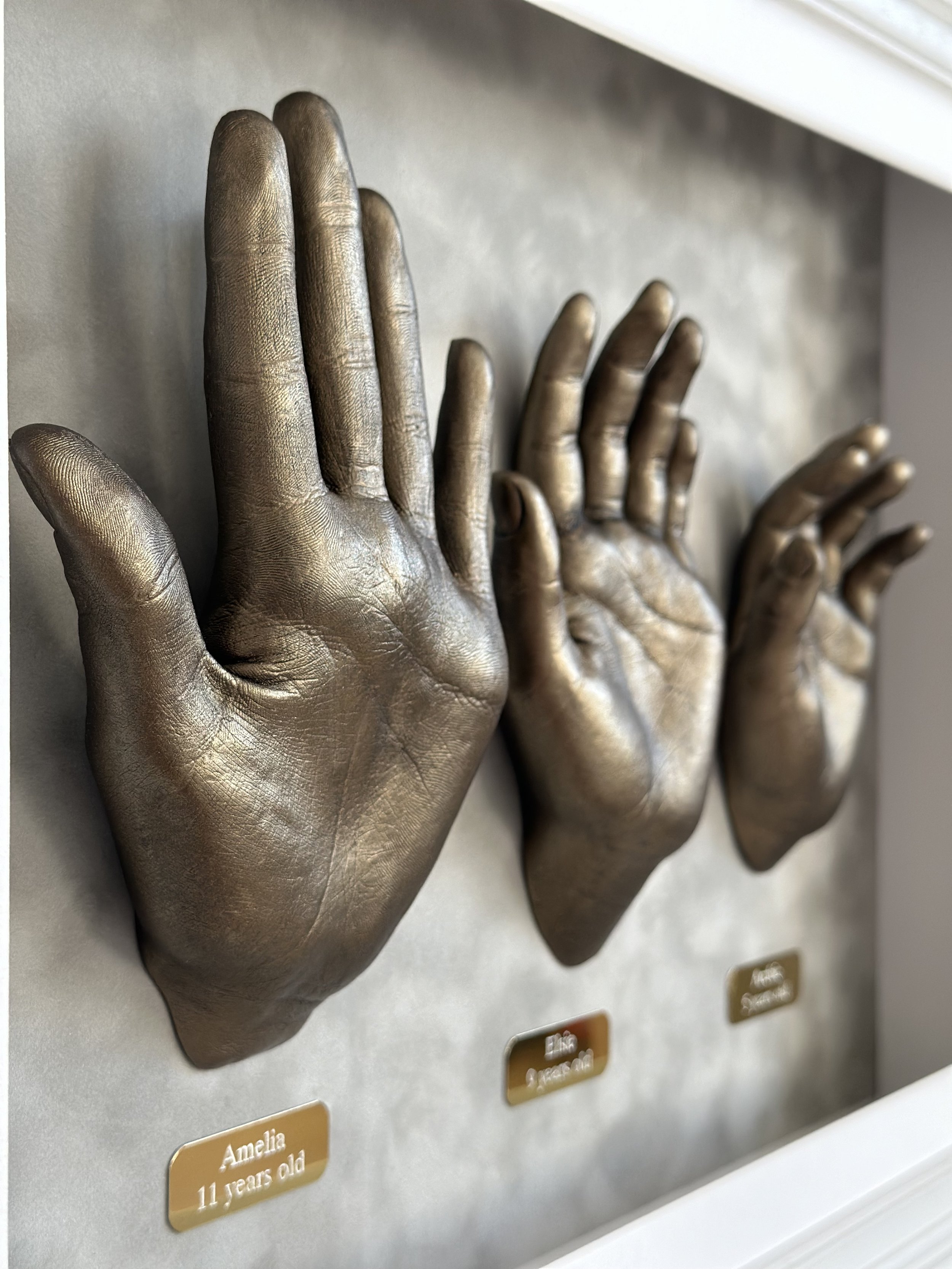 Bronze-colored plaster hand sculptures displayed on a wall with engraved nameplates showing ages.