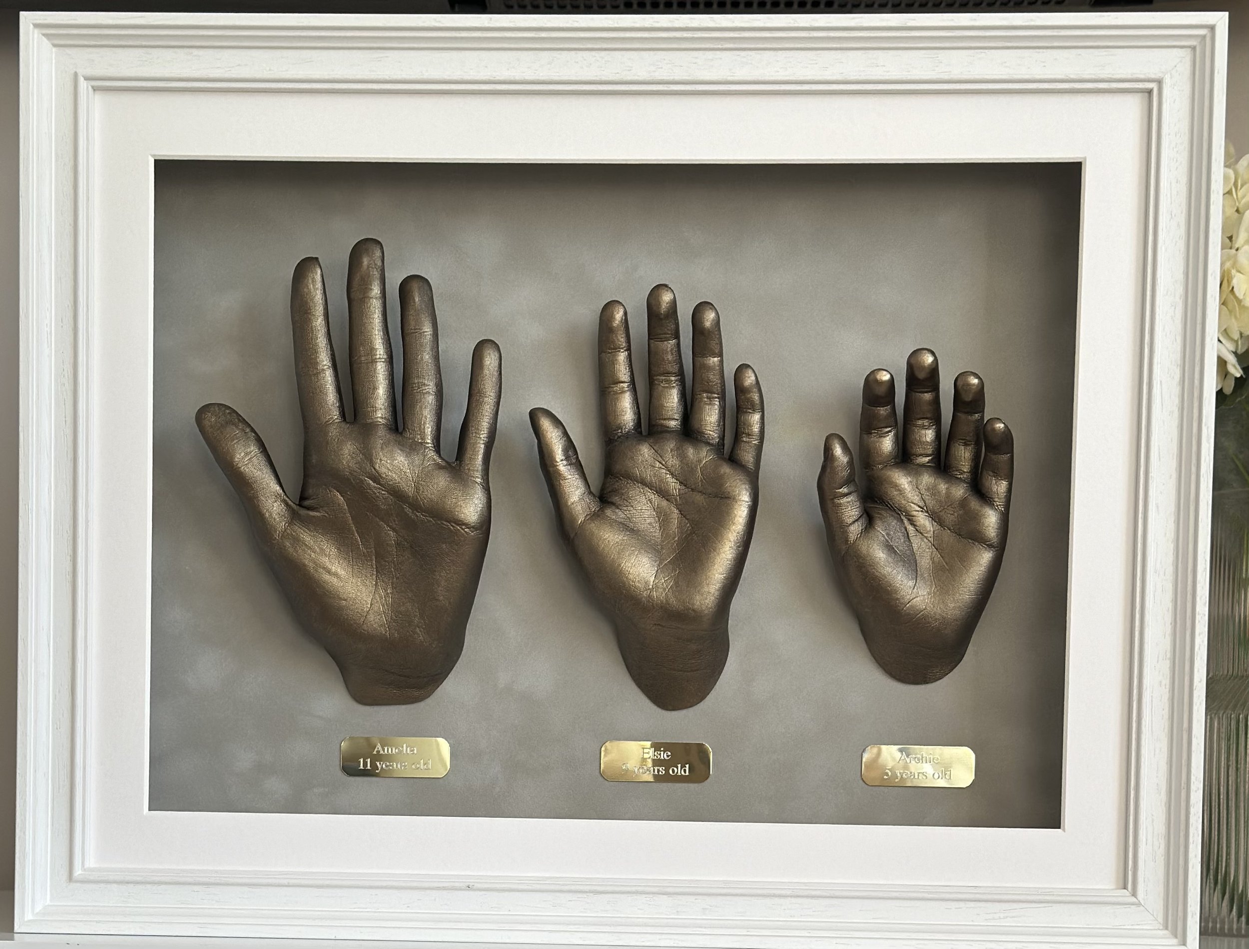 Framed display with three bronze hand casts labeled for different ages, set against a gray background.