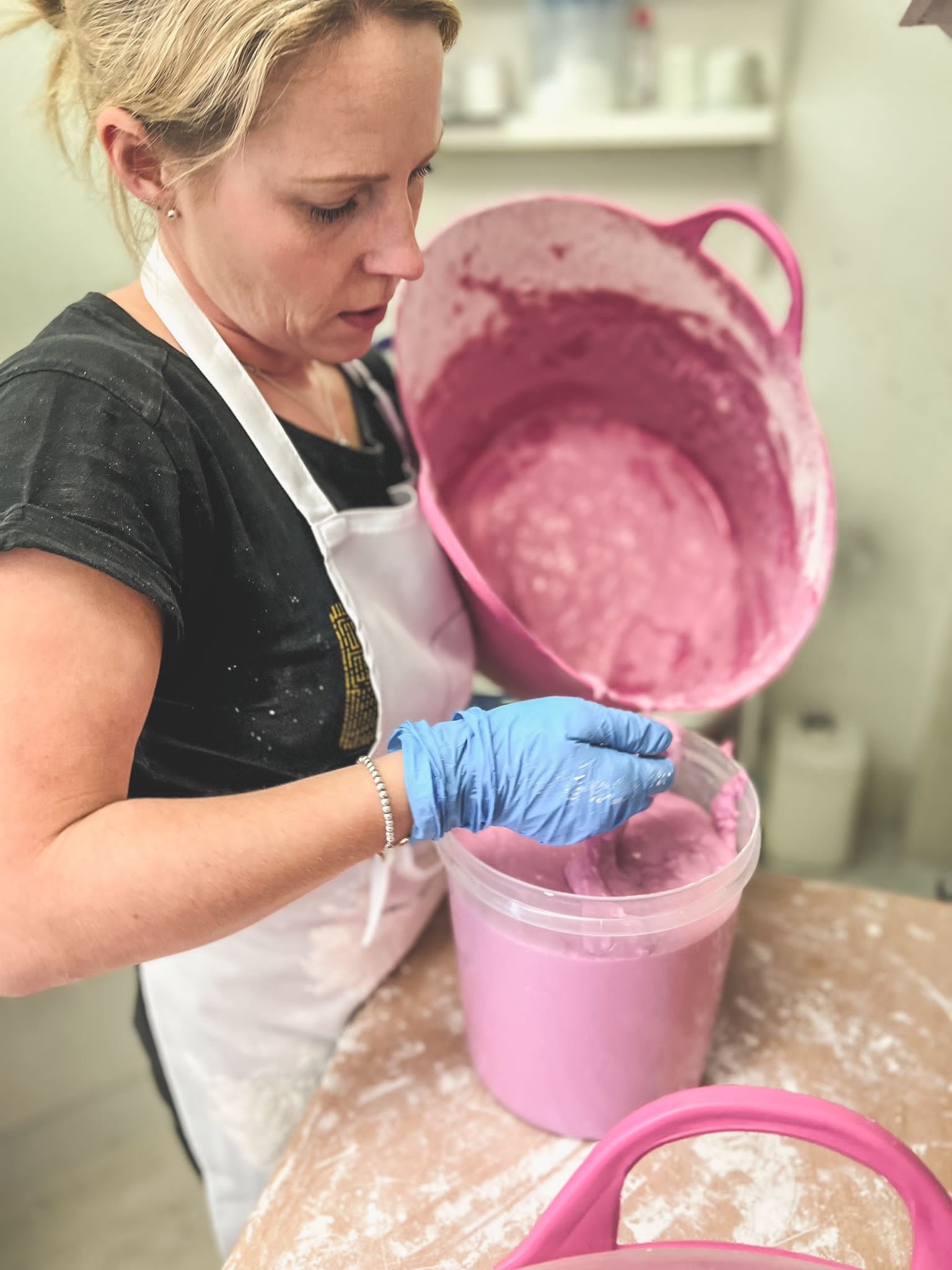 A woman wearing a black shirt and white apron is pouring pink mixture from a container into a bucket. She is wearing blue gloves and is focused on her task. The setting appears to be a kitchen or workshop with visible shelves and supplies in the back