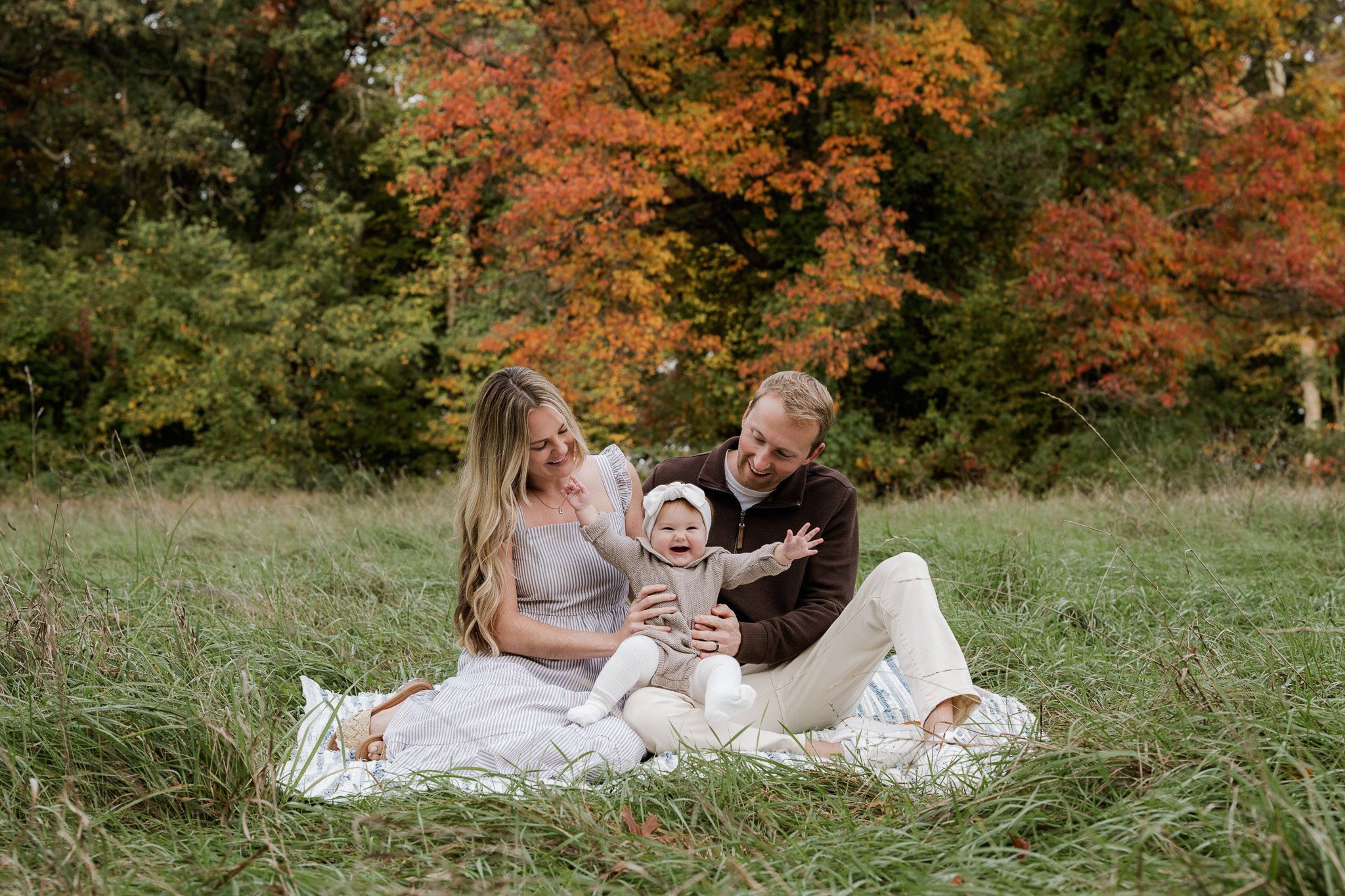Metro Detroit photographer captures two parents in a park during fall, enjoying time with their 8 month baby. In a park with Fall foliage.