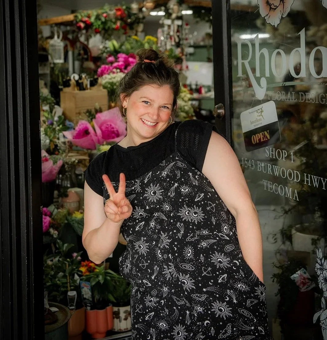 Woman smiling and making a peace sign at the entrance of a flower shop, surrounded by colorful flowers and greenery.