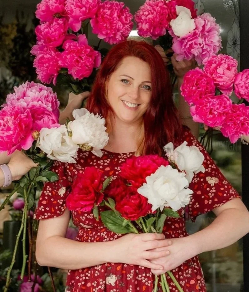 A woman with red hair smiling and holding a bouquet of white and red peonies, surrounded by pink peonies.