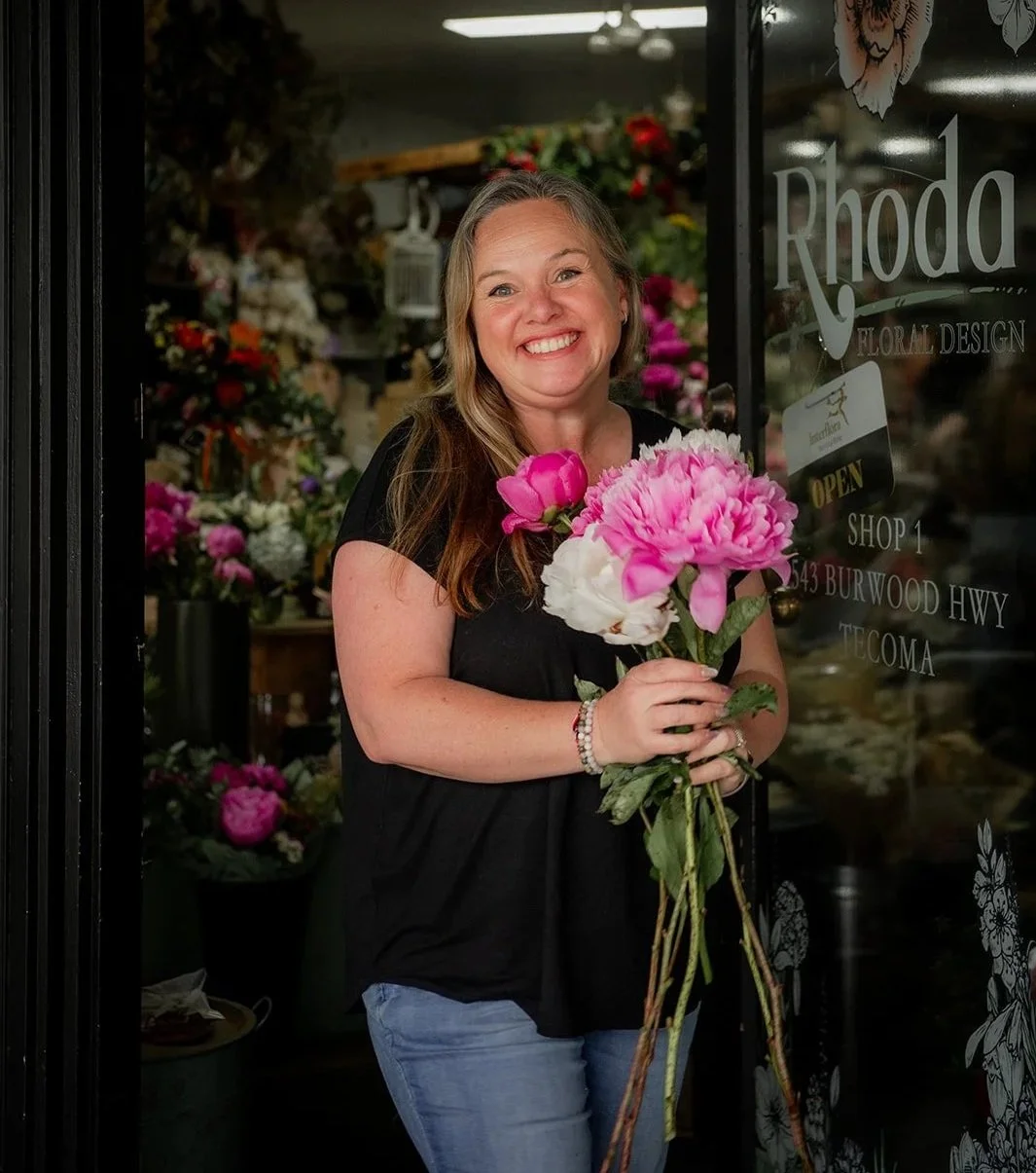 A smiling woman holding pink and white peonies in front of a flower shop window.