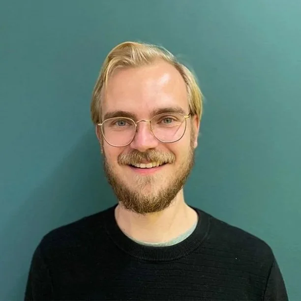 A young man with blonde hair, glasses, and a beard smiling against a teal wall.