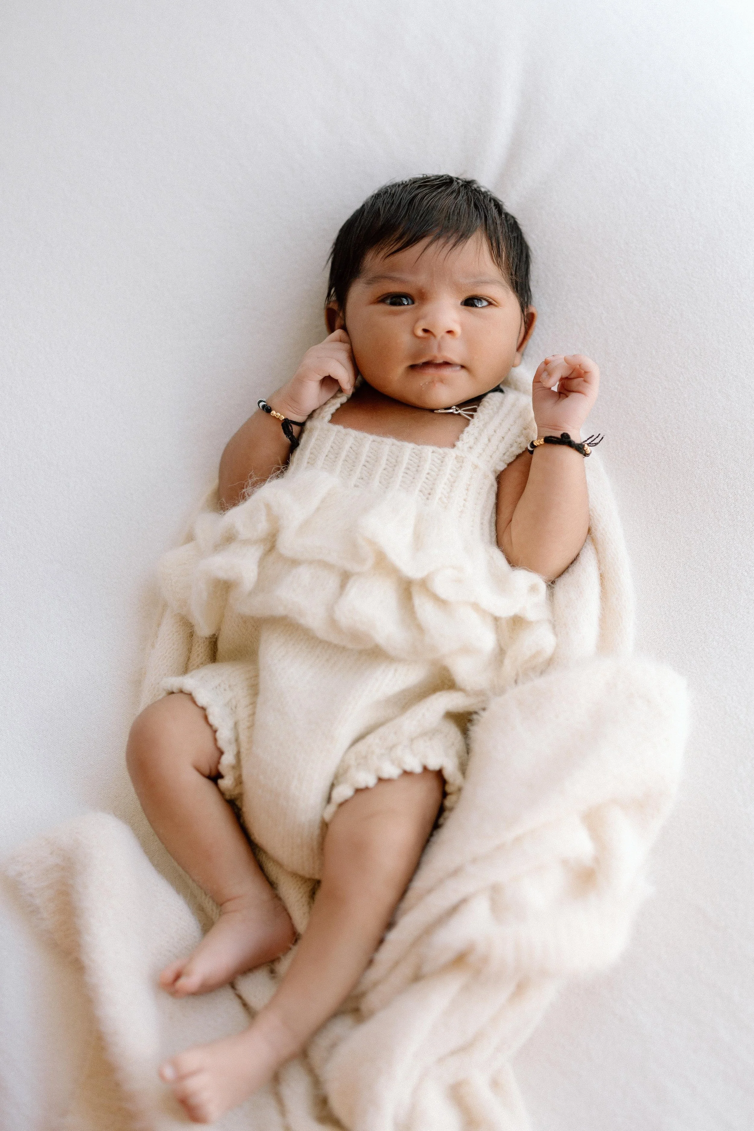 A baby lying on a white blanket, wearing a cream-colored knitted romper with ruffles, looking directly at the camera.