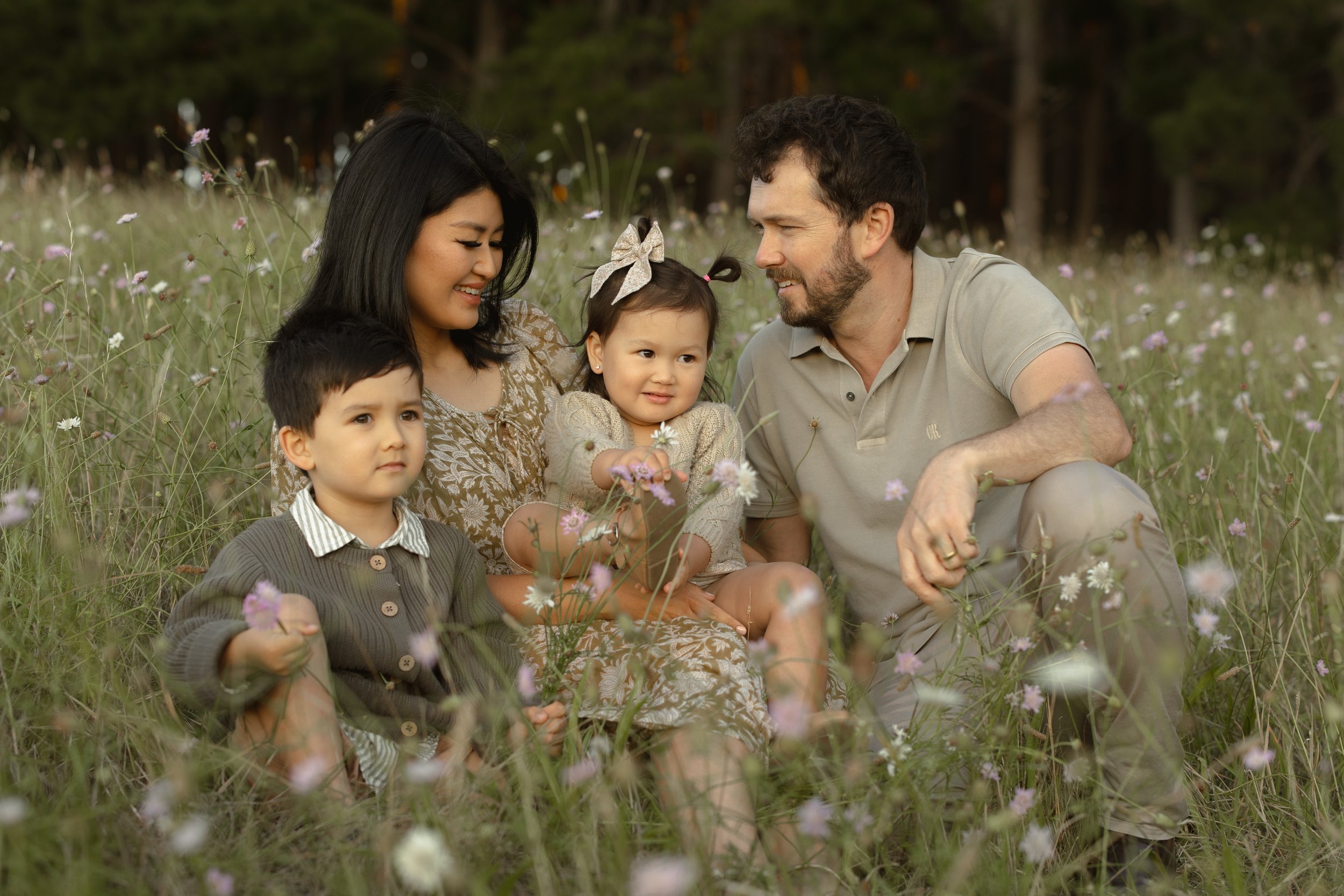 A family of four sitting in a grassy field with wildflowers, smiling and enjoying each other's company.