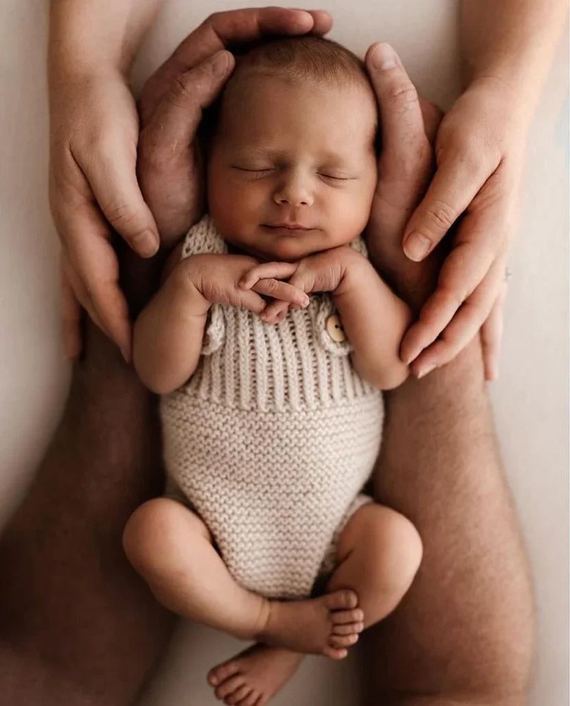 A sleeping newborn baby wrapped in cream knitted blankets and wearing a matching bonnet, lying on a soft white surface.