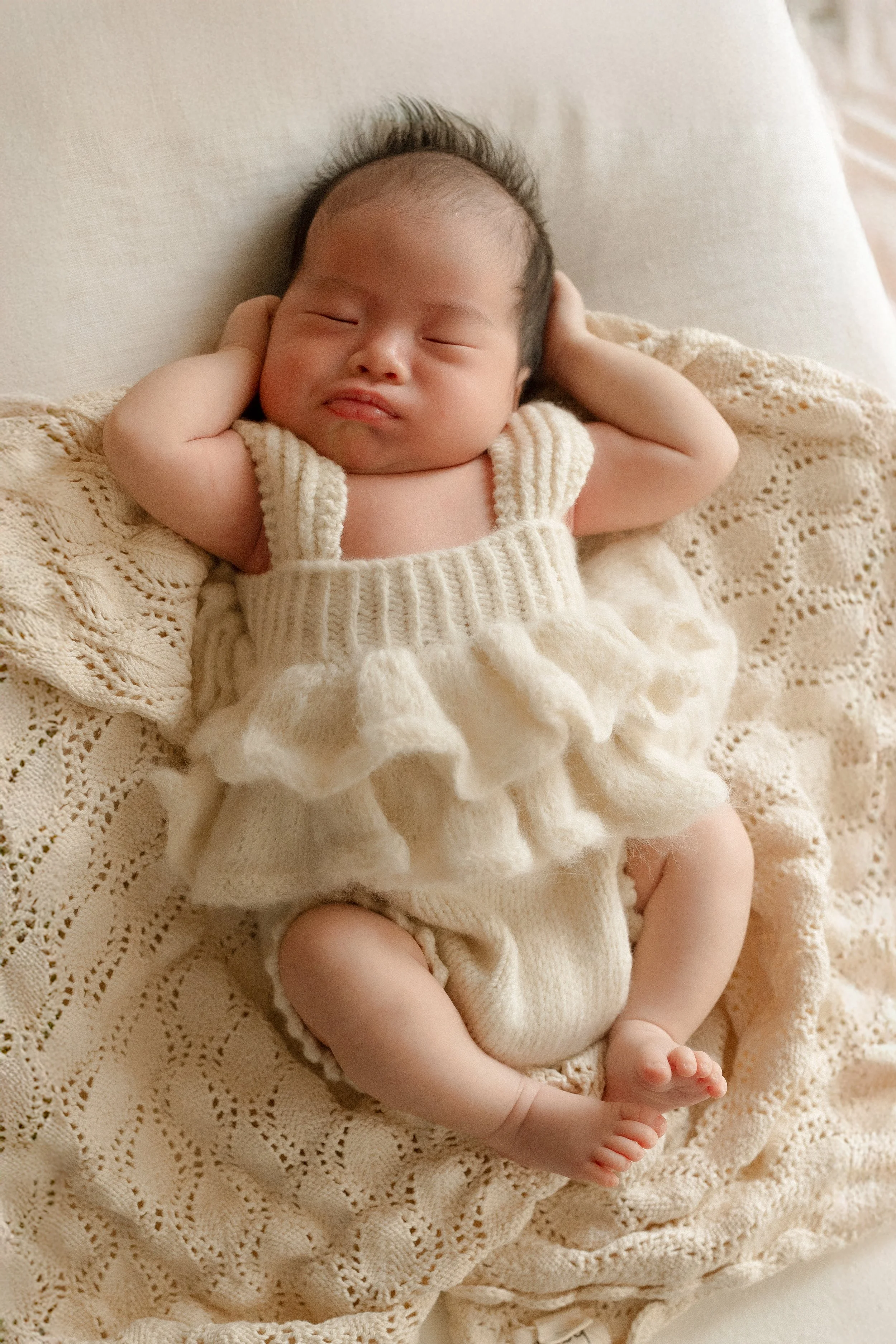A baby lying on a white blanket, wearing a cream-colored knitted romper with ruffles, looking directly at the camera.