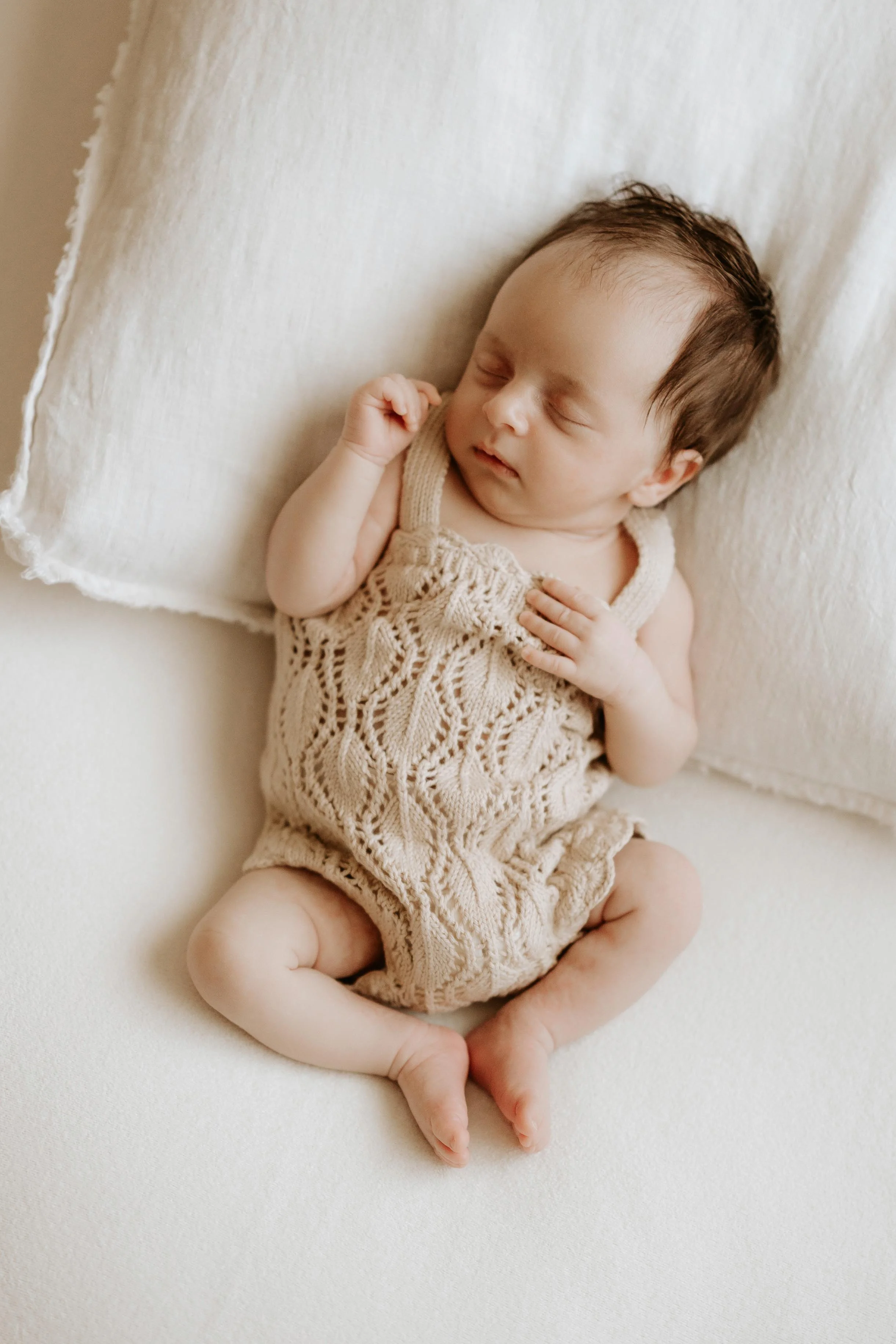 A sleeping baby with dark hair, lying on a white pillow, wearing a beige knitted outfit.
