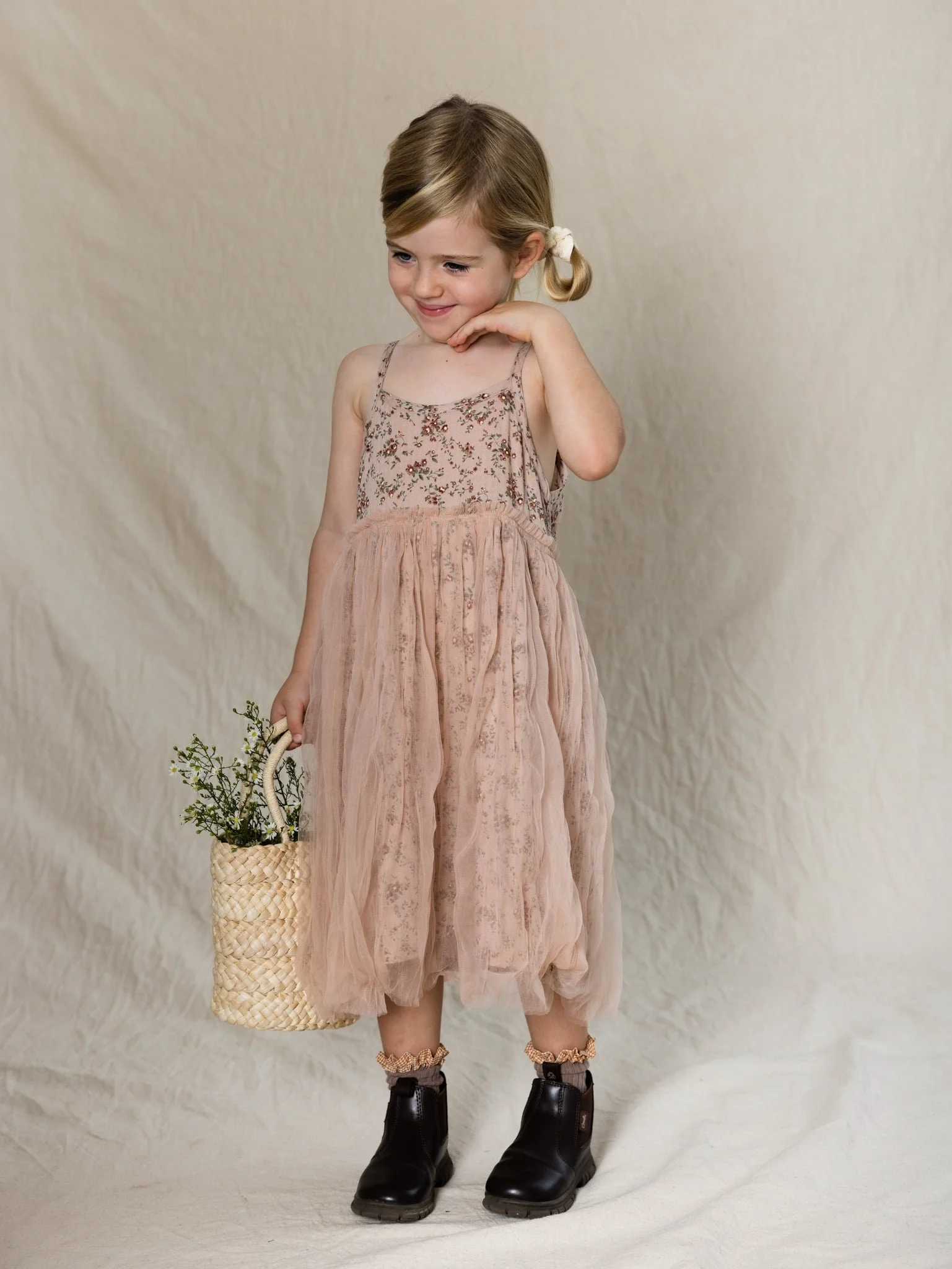 Young girl in a pink floral dress and black boots holding a basket with greenery, standing against a plain beige background, smiling and looking down.