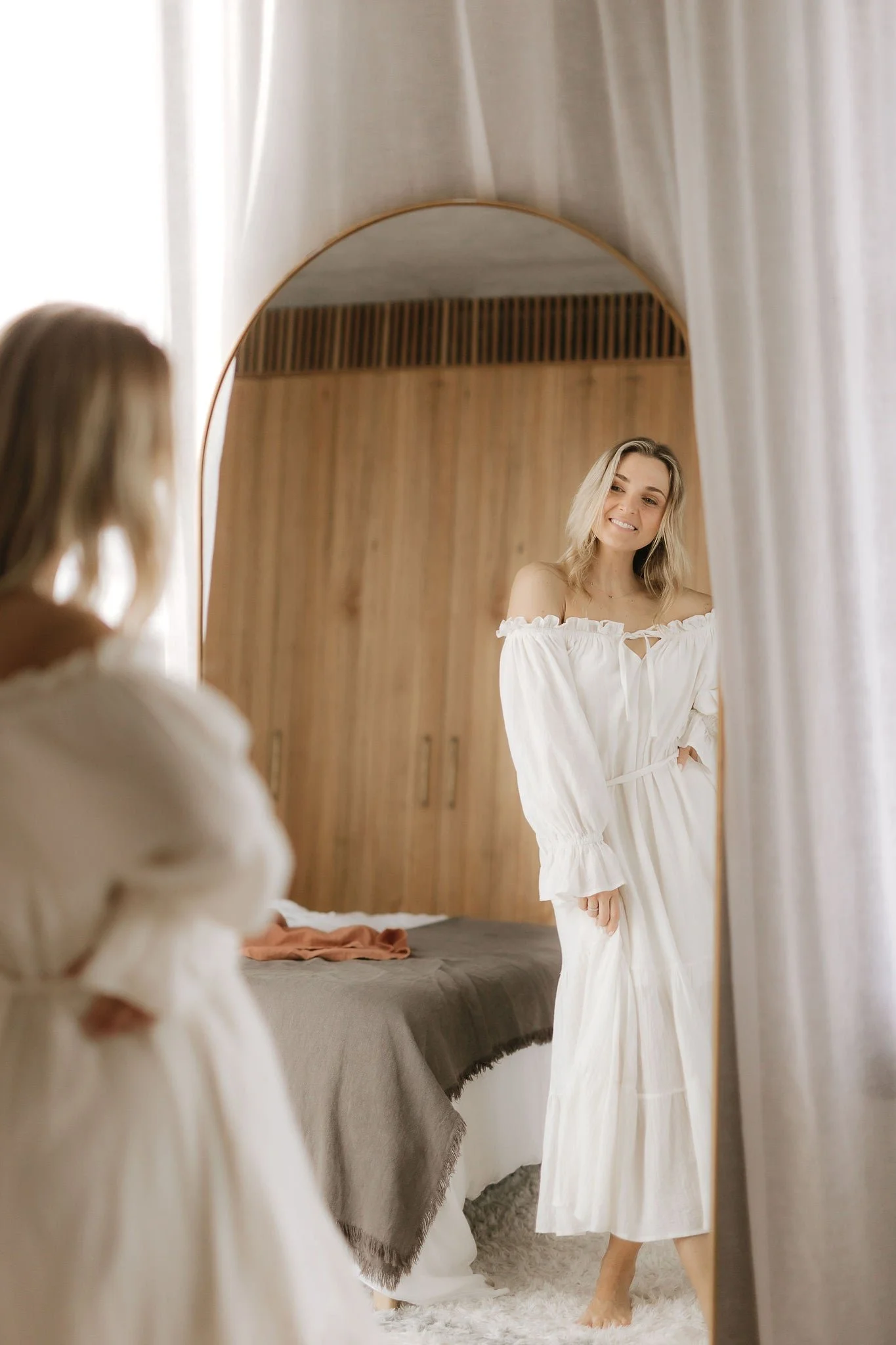 A woman in a white dress smiling as she looks into a mirror. A girl in a similar dress is blurred in the foreground, with a bedroom in the background.