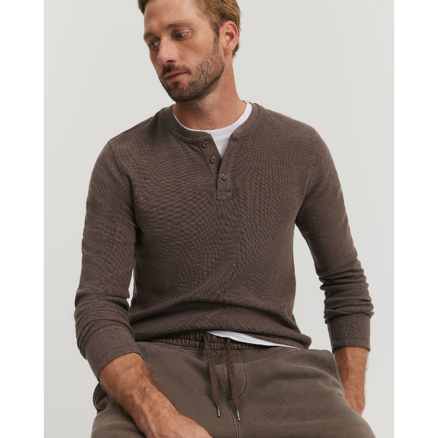 Man wearing a brown long-sleeve henley shirt and khaki sweatpants sits against a plain background.