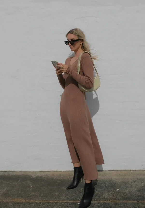 Woman with blonde hair wearing black sunglasses, a brown jumpsuit, and black ankle boots, holding a phone, standing against a white wall, with a beige handbag over her shoulder.