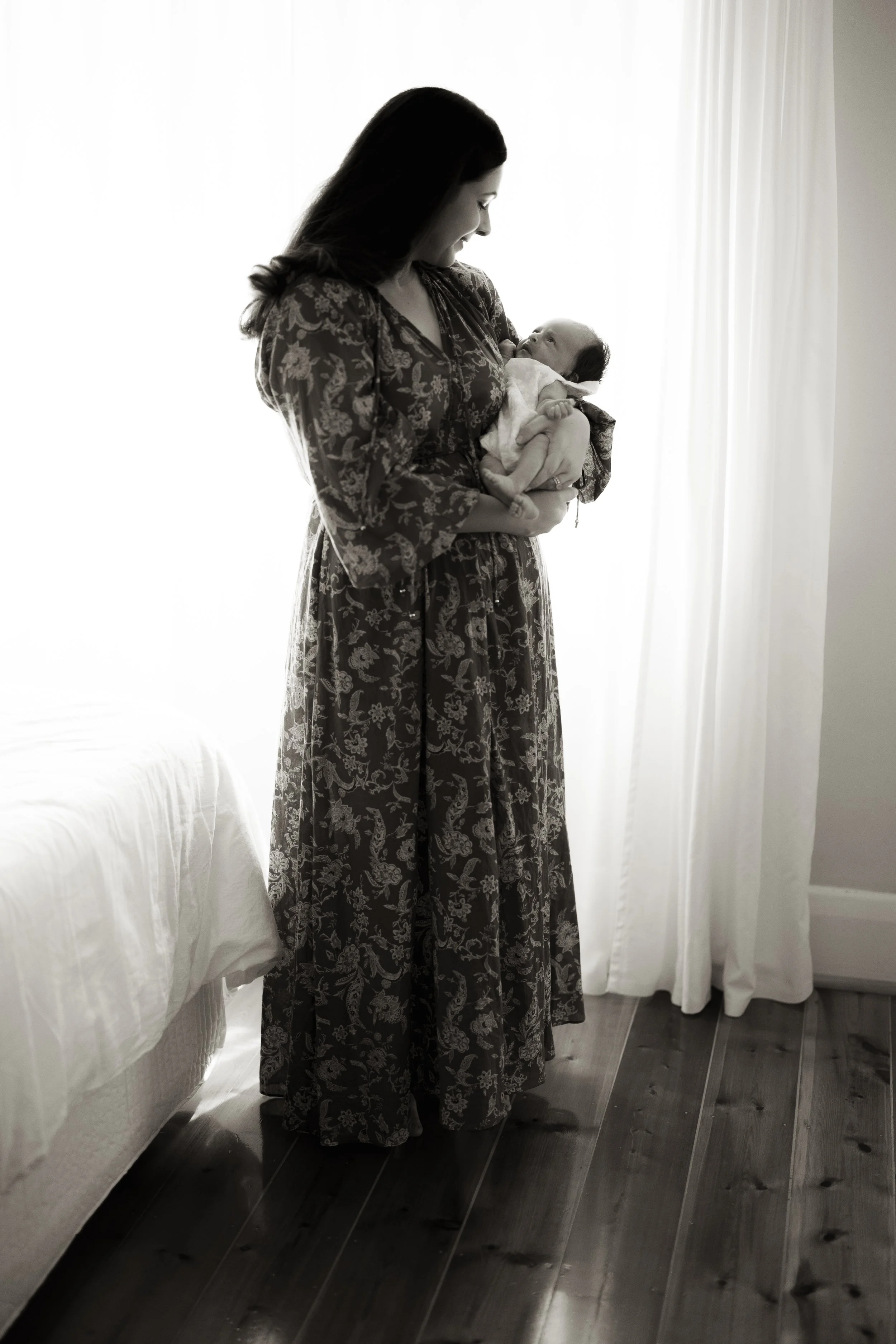 A woman in a floral dress holding a newborn baby near a window with white curtains, black and white photo.