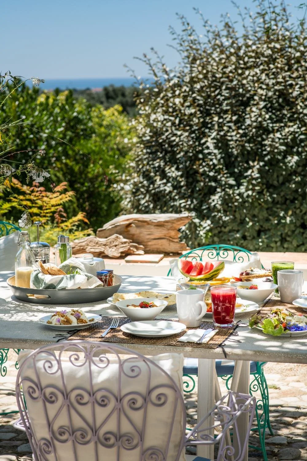 Outdoor breakfast table set with dishes, beverages, and fruit, surrounded by green bushes and trees, with a view of the sky and ocean in the background.