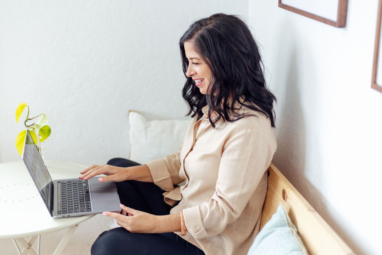 Charmaine Renee, rest coach and breathwork practitioner,  sitting on a wooden bench, looking at a laptop on a small white table, with a potted plant nearby and framed pictures on the wall.